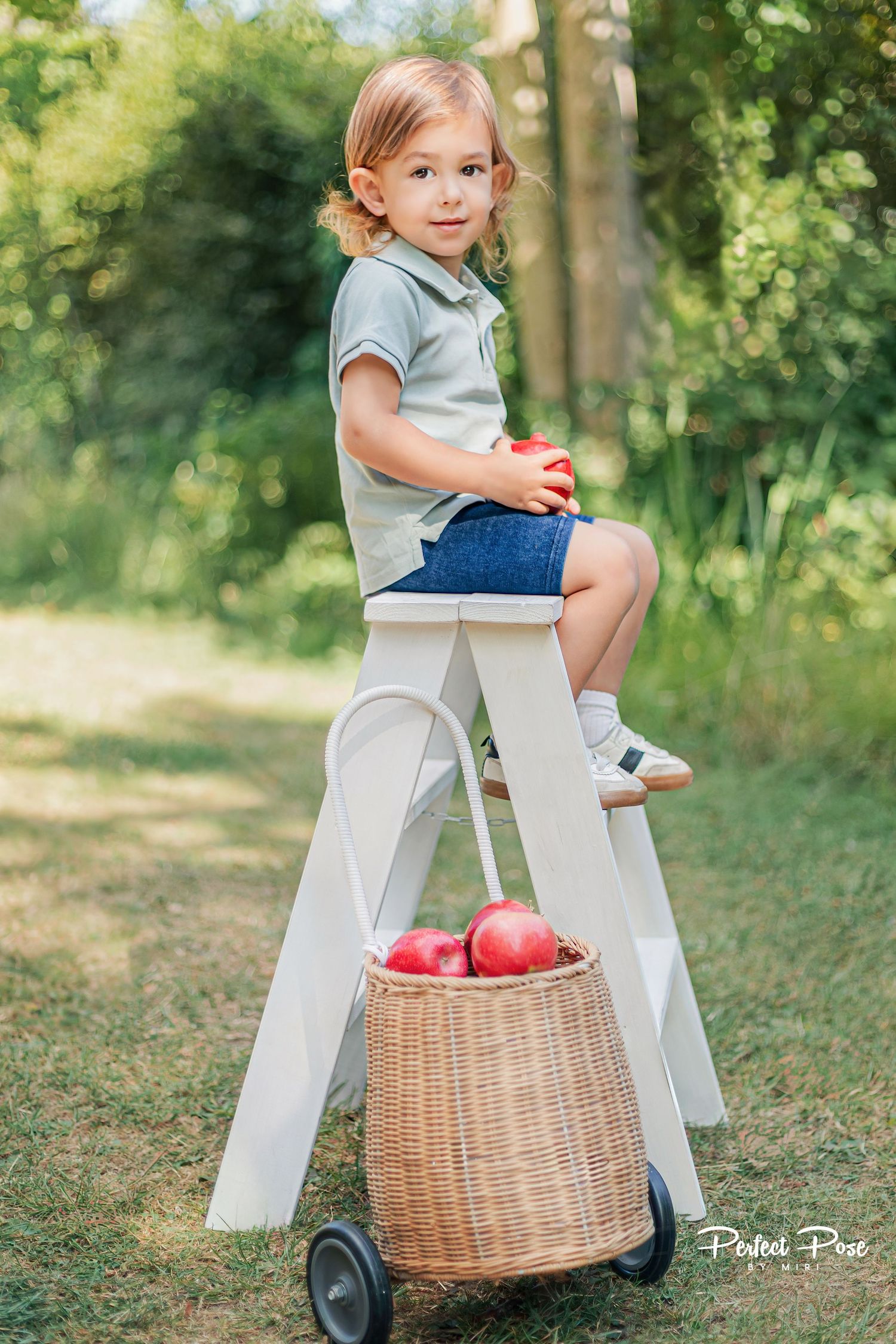 A young child sits on a white step stool holding a red apple, with a wicker basket of apples nearby in a lush garden.