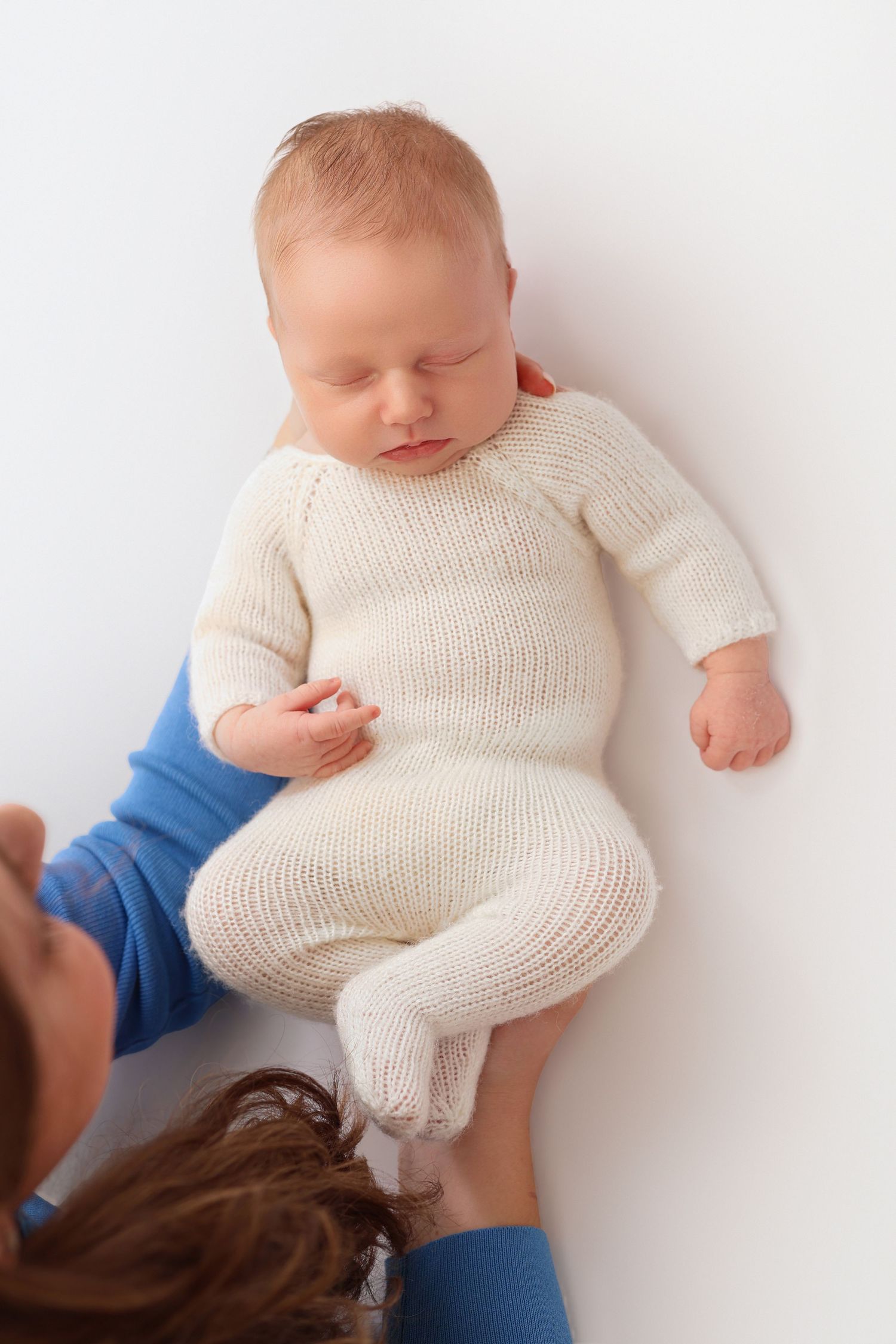 Sleeping newborn baby in white knit romper held gently by parent hands on white background.