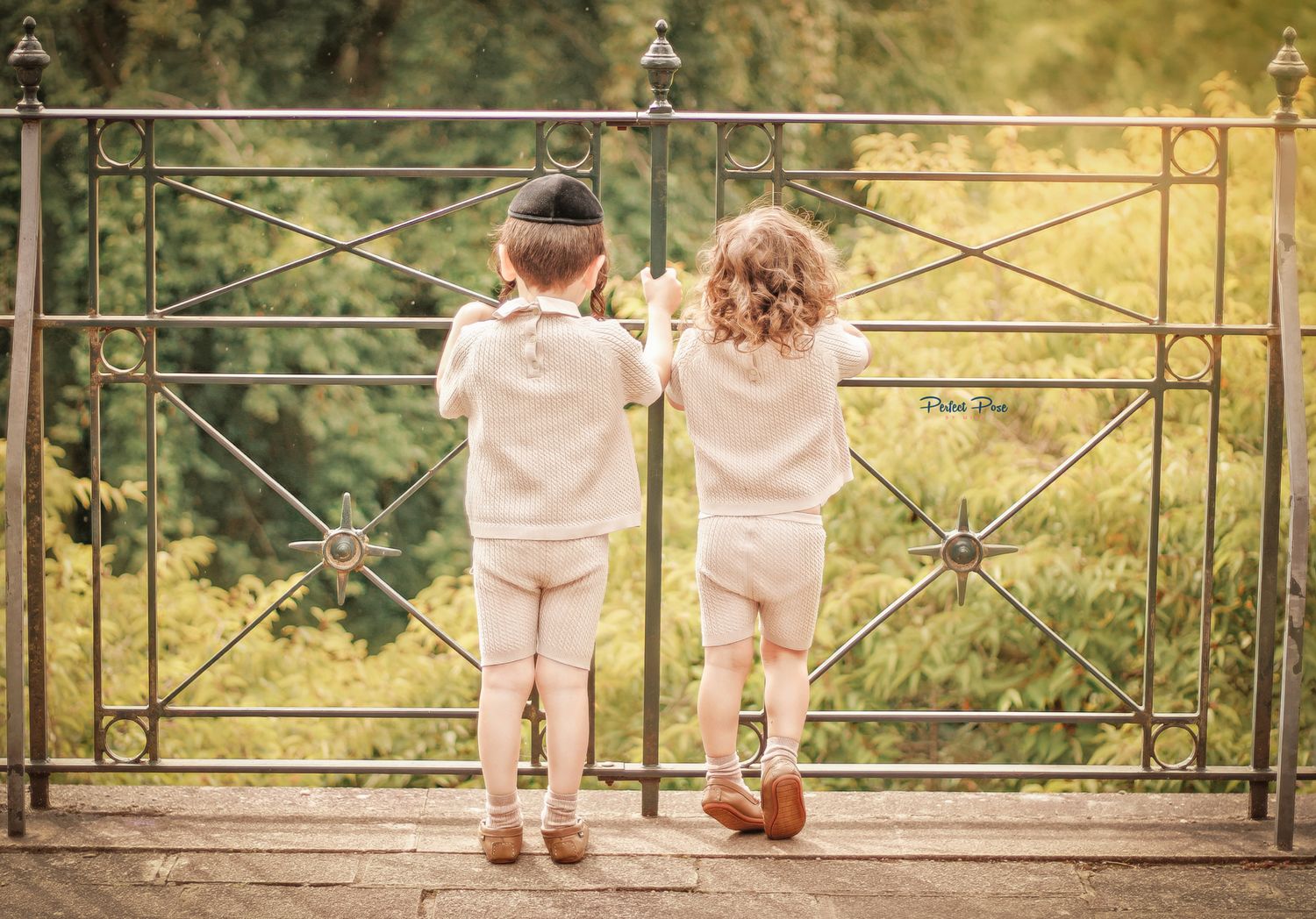 Two young children in matching cream outfits stand holding hands at an ornate iron gate, gazing into a lush green garden.