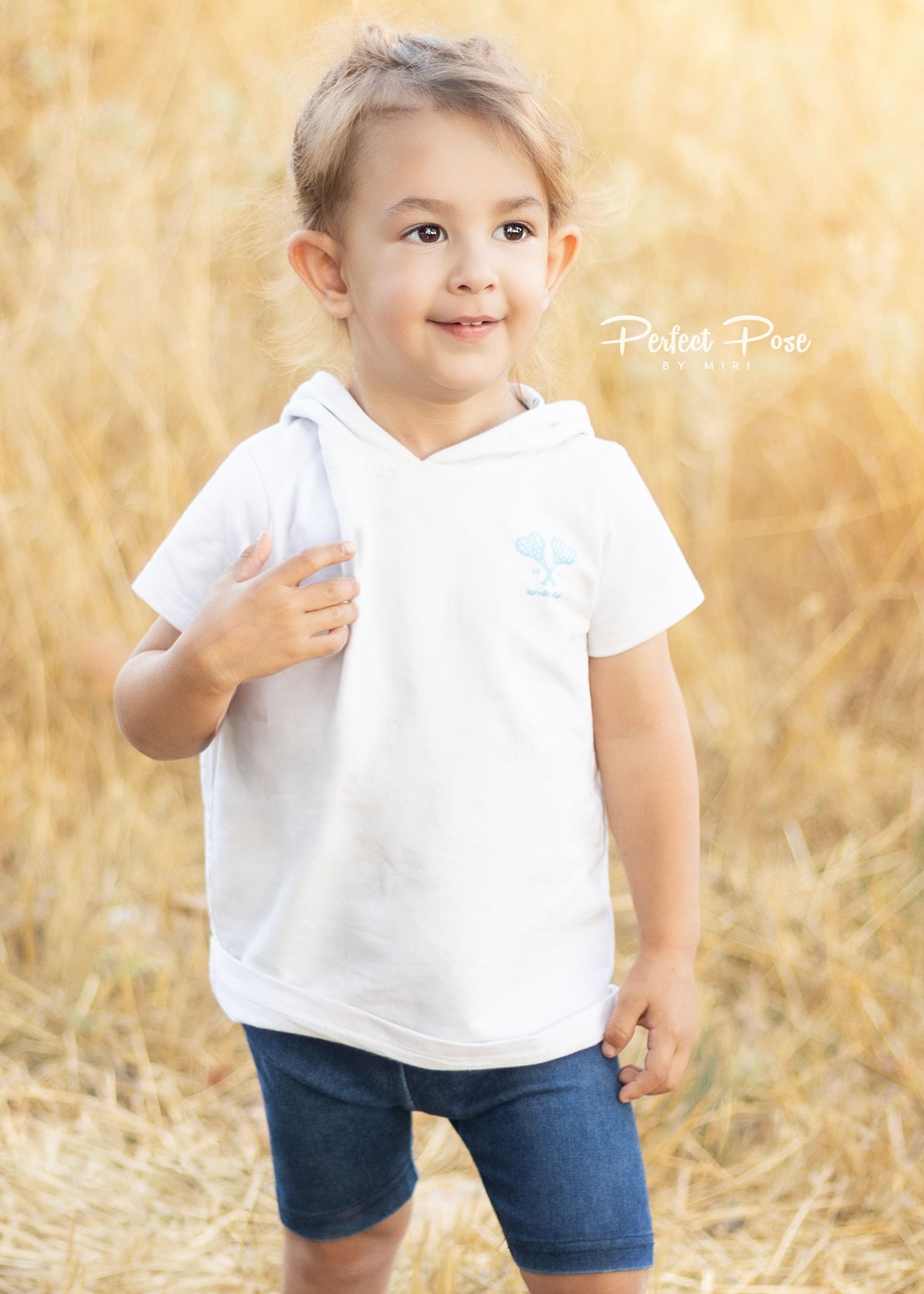 Smiling toddler in white hooded tee and denim shorts posing in golden field, professional children's portrait photography.