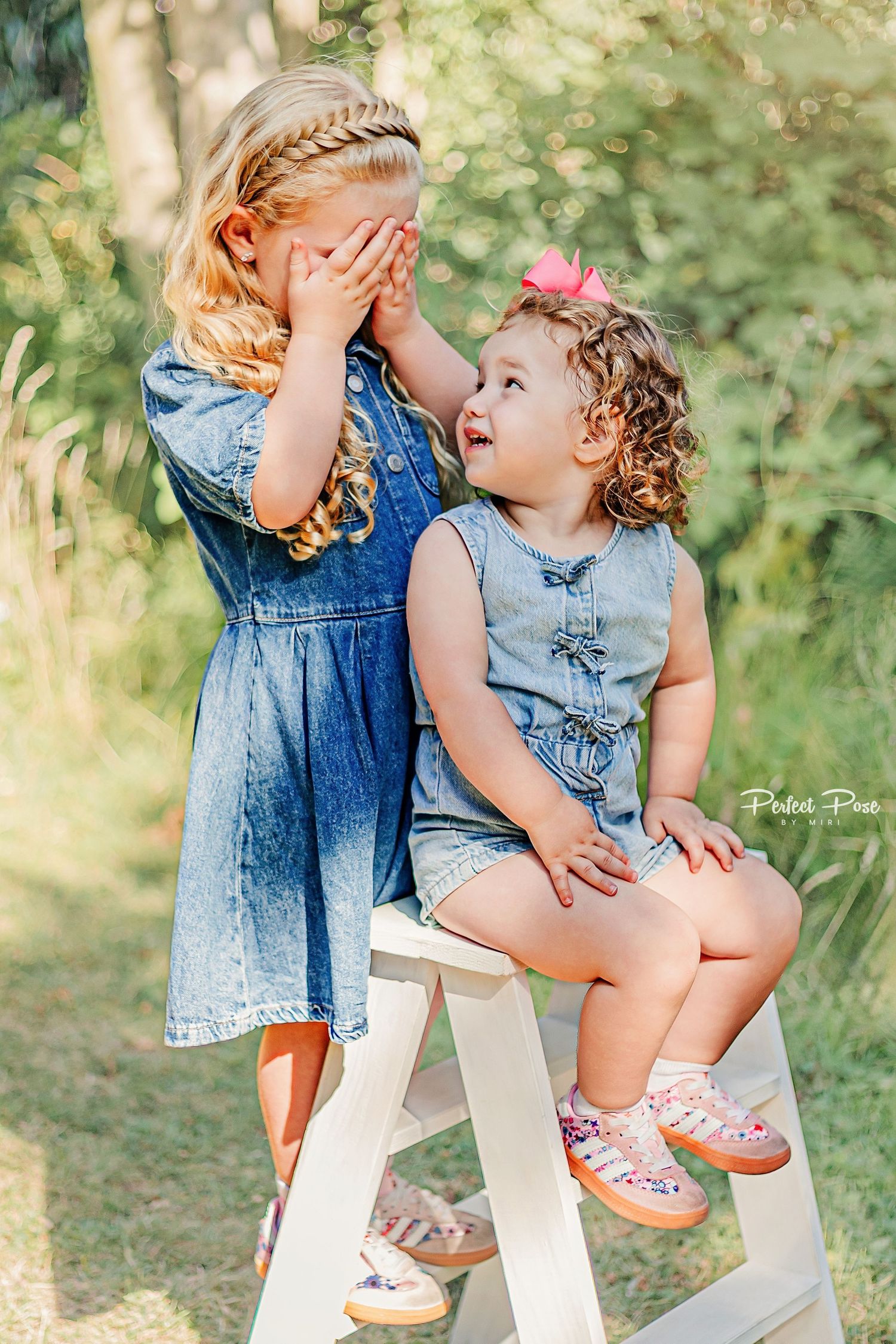 Two young sisters in matching denim outfits share a playful moment outdoors, one covering her eyes while the other laughs.