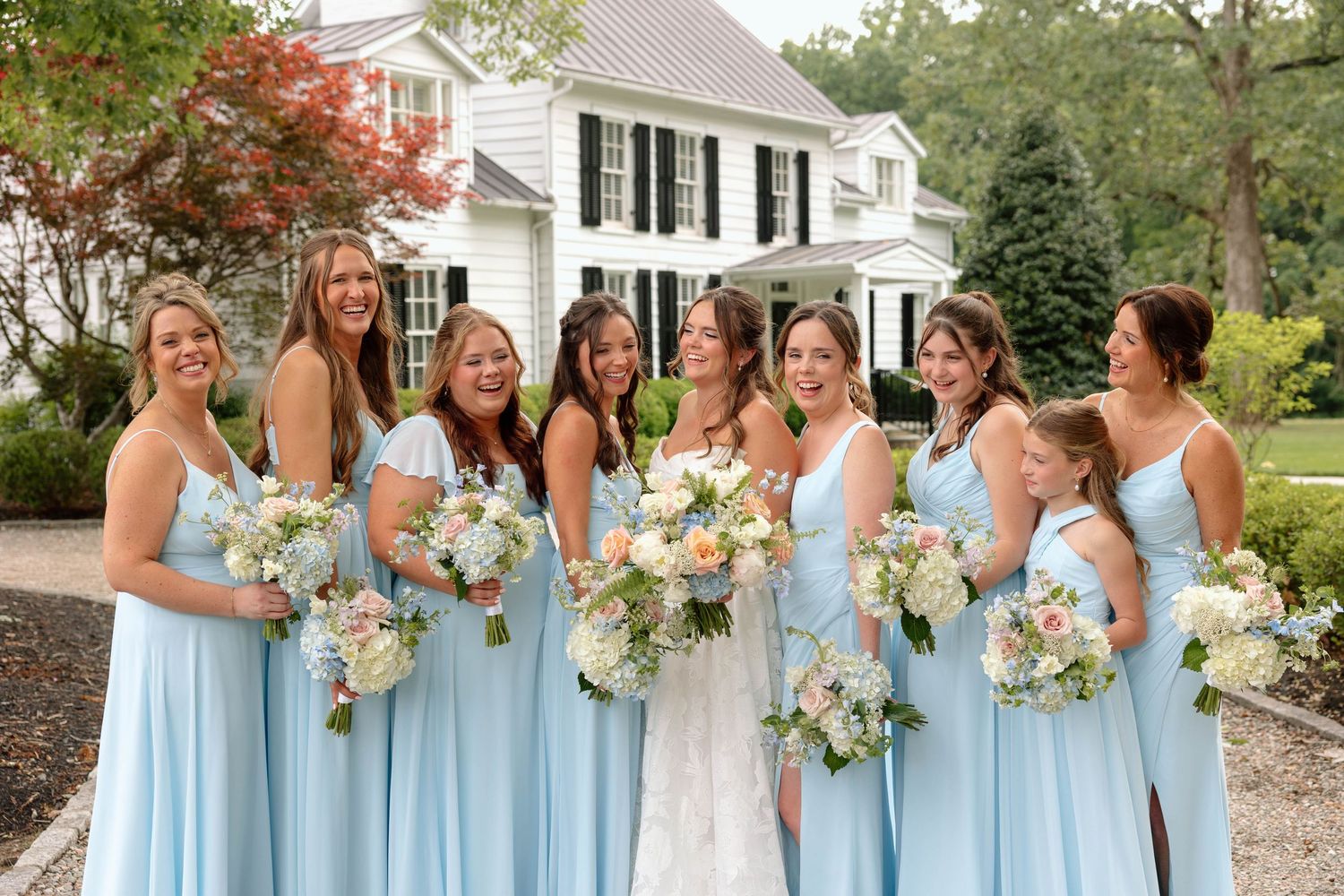 Bride and her bridesmaids in light blue standing in a line laughing together holding their bouquets at various heights.