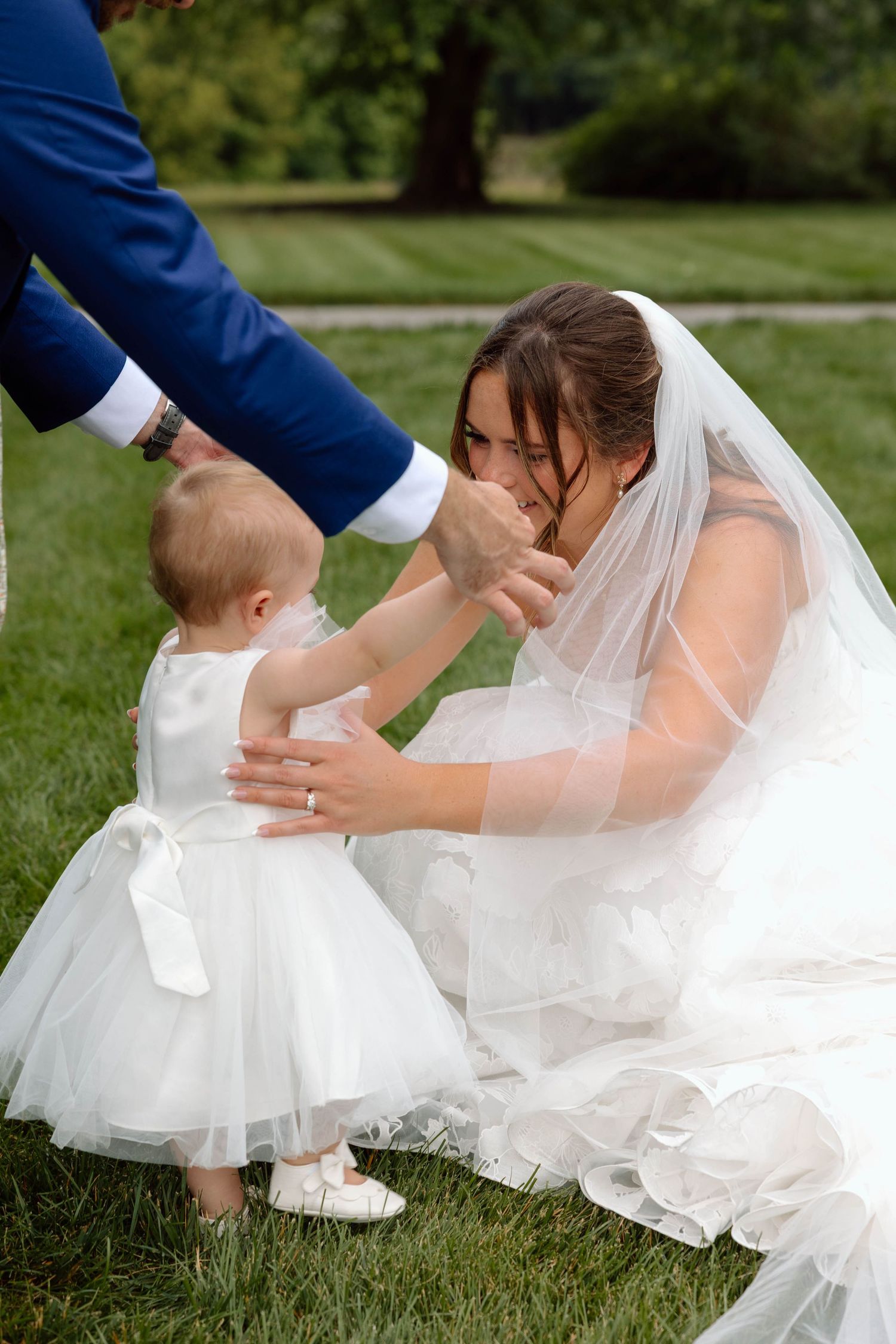Bride wearing a long veil squatting down with a toddler walking towards her.