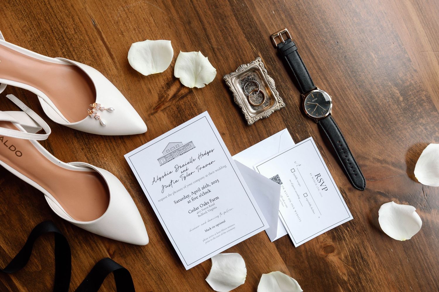 White heels with gold jewelry and a white invitation suite on wood. Surrounded by white rose petals with a men's black leather watch.