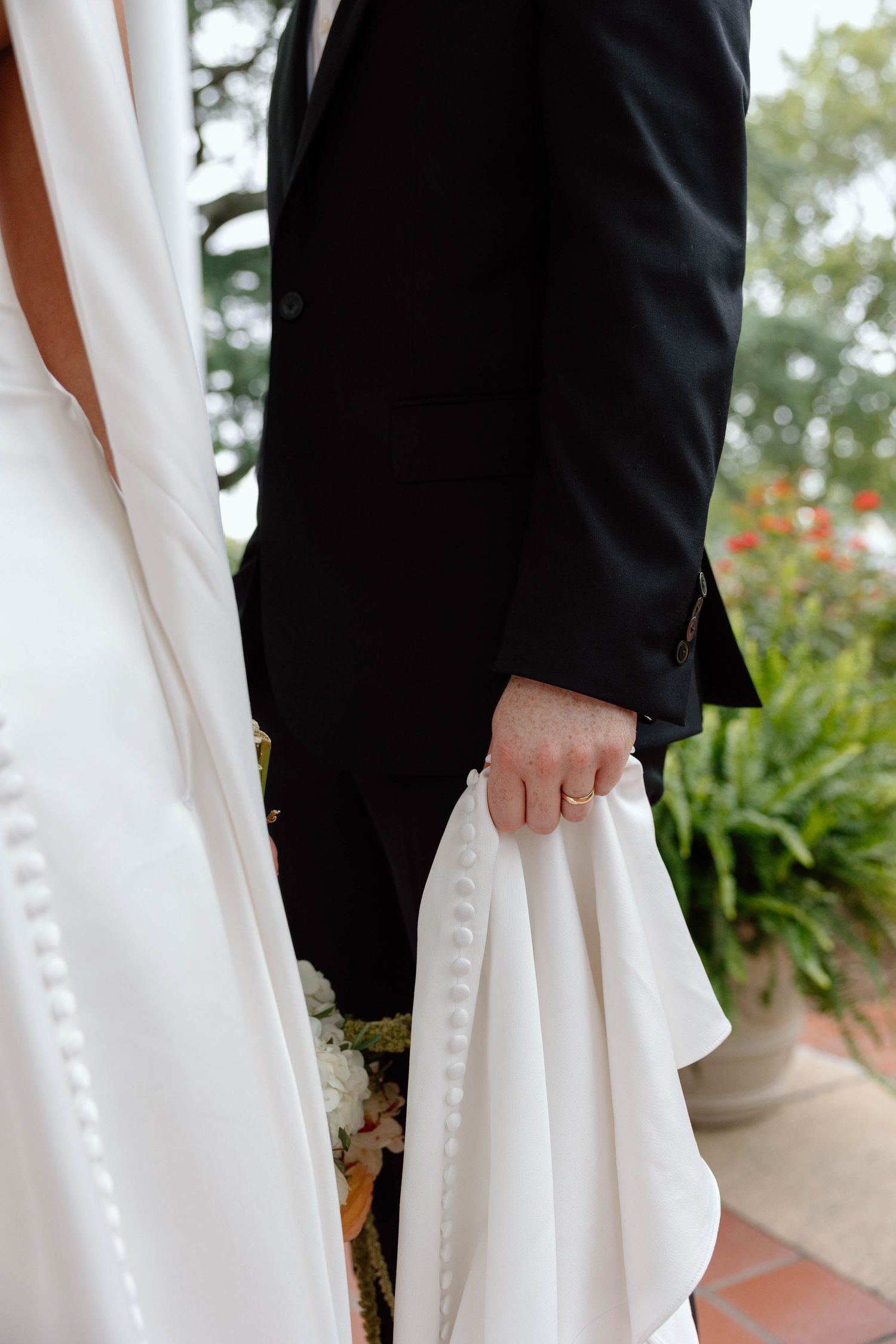 Groom holding the train of bride's dress with his left hand. Simple gold wedding band showing.