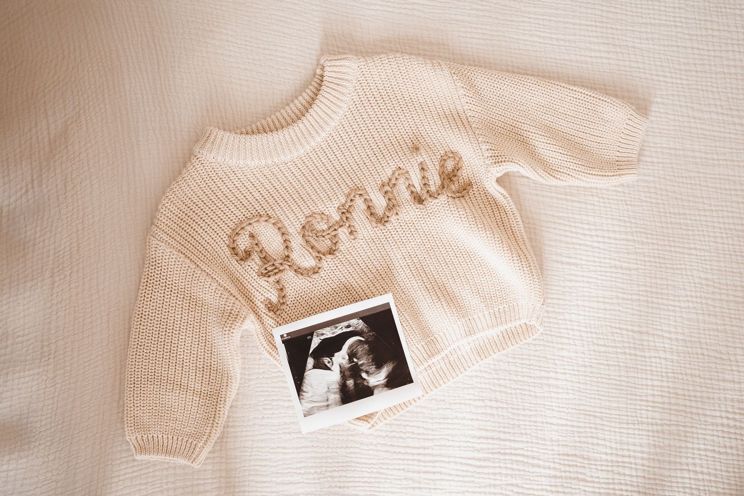 Cream knit baby sweater with embroidered name Ronnie displayed flat with black and white ultrasound scan photo.