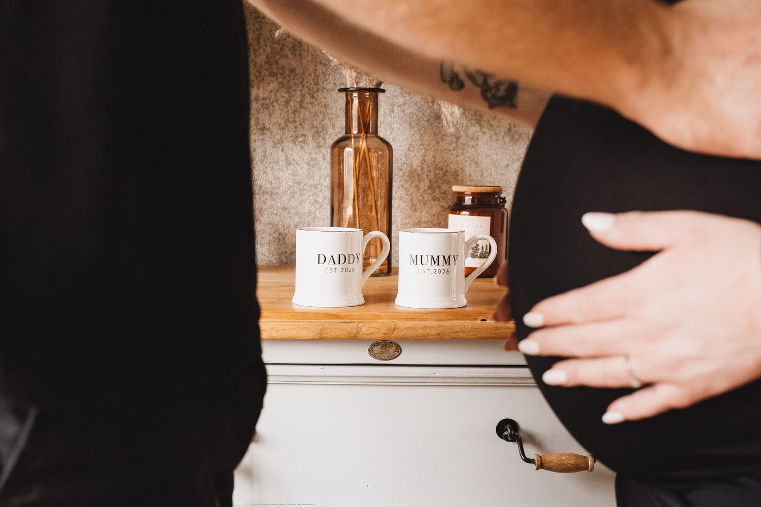 A pregnant belly beside a kitchen shelf displaying Daddy and Mummy mugs on a wooden tray.