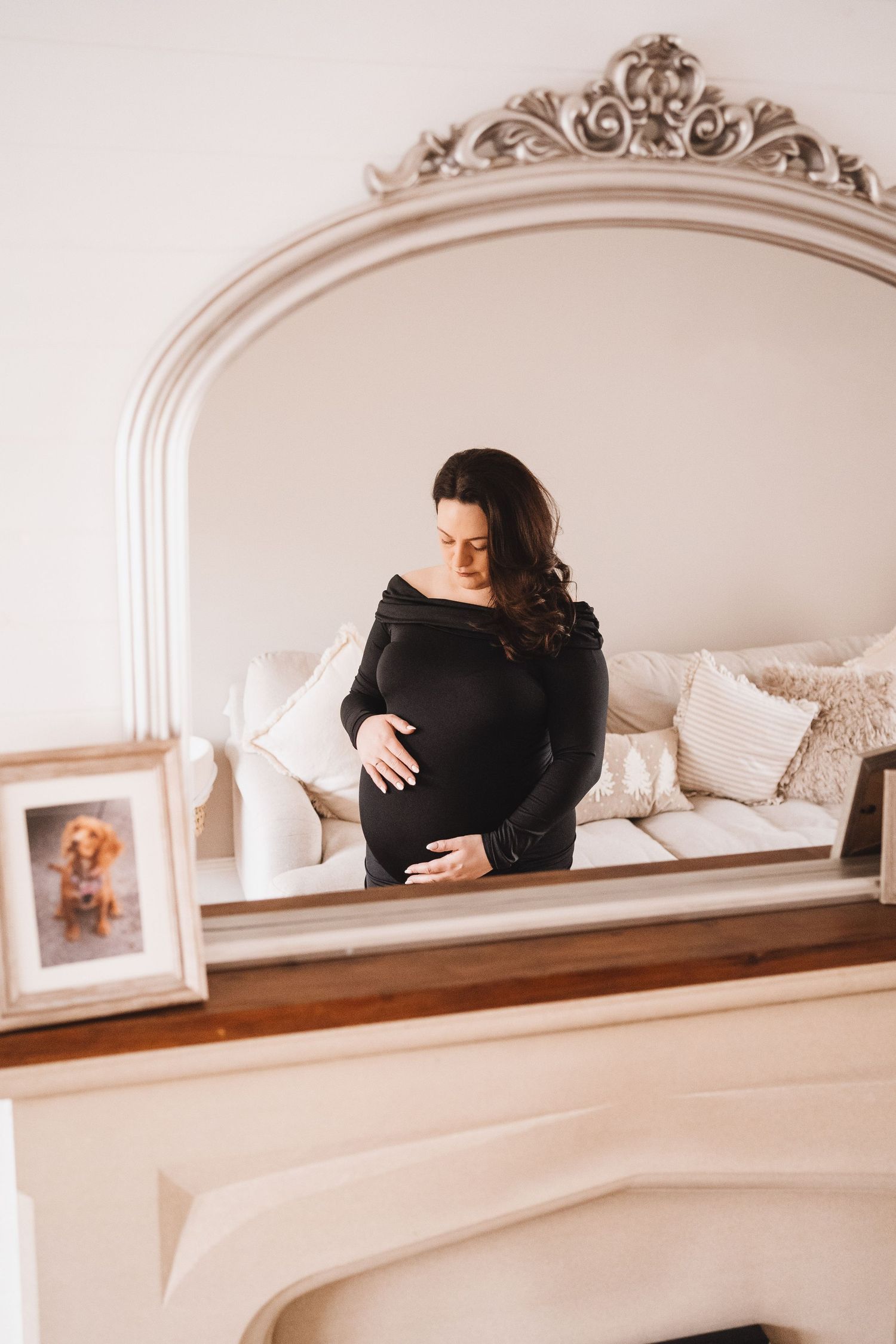 A pregnant woman in a black dress cradles her bump while reflected in an ornate silver mirror.