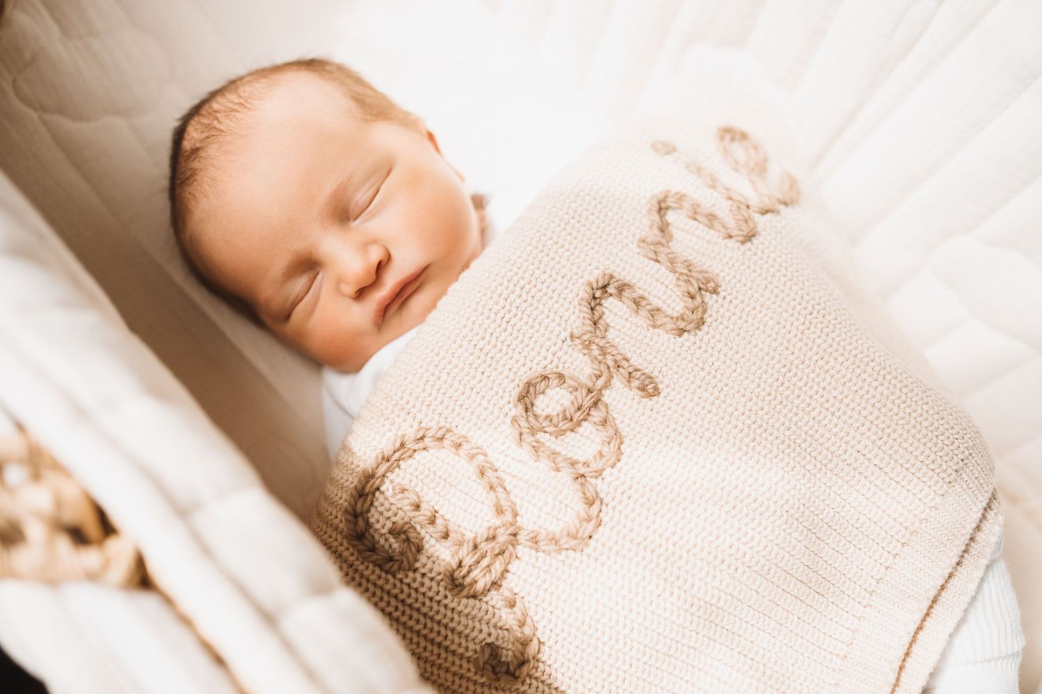 Peaceful sleeping newborn baby wrapped in personalized cream knitted blanket with embroidered name in white crib.