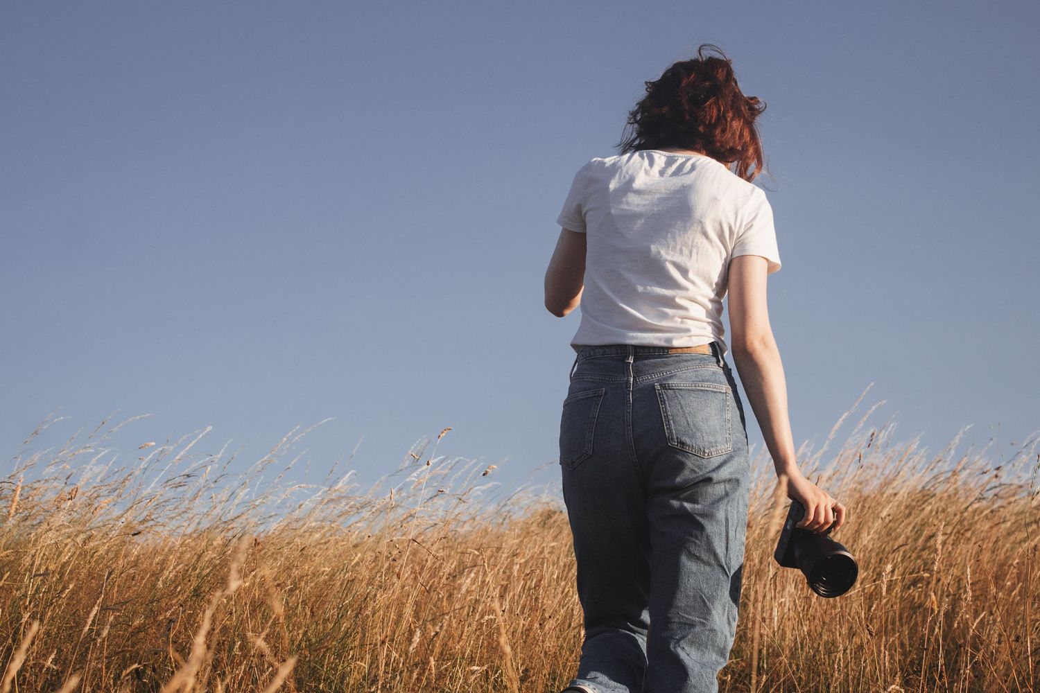 Woman in white t-shirt and jeans walking through golden wheat field holding camera from behind.