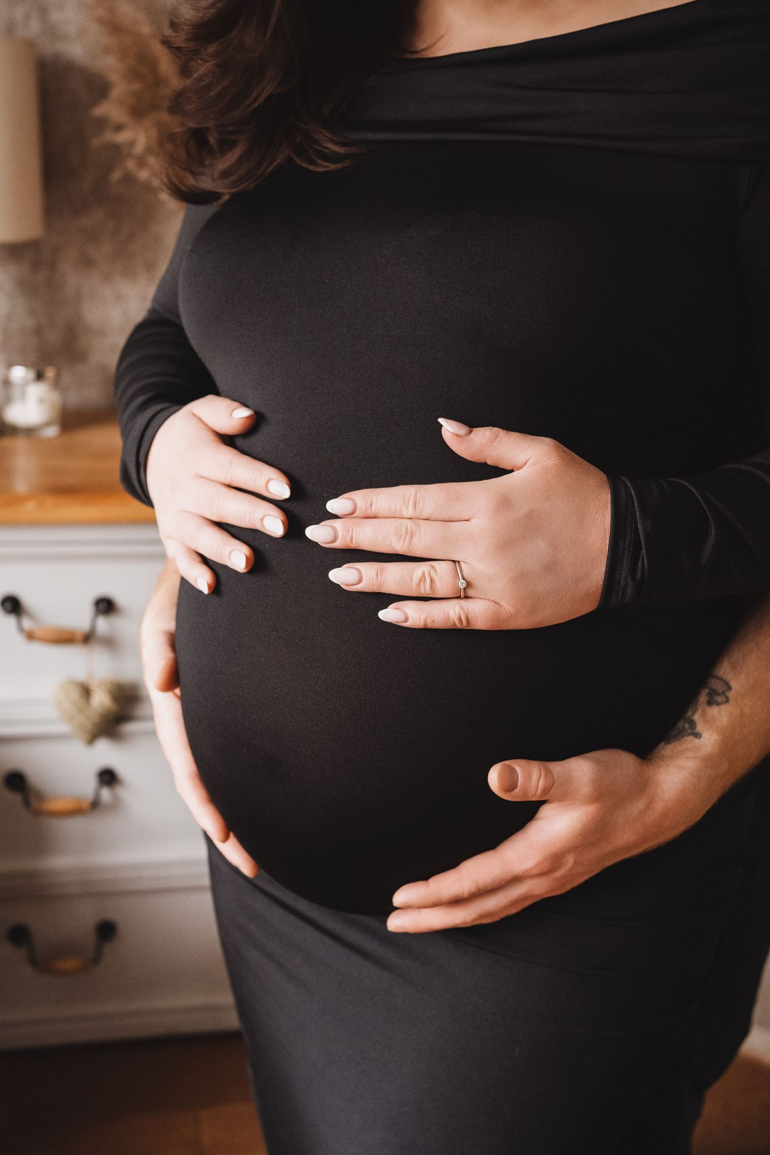Close-up of a pregnant woman in a black dress with her partner's hands cradling her baby bump.
