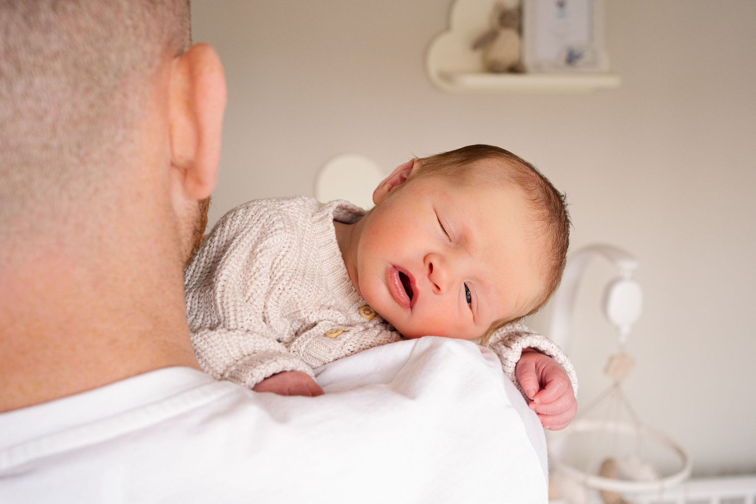 Father holding a sleeping newborn baby dressed in a beige knit outfit against his shoulder.