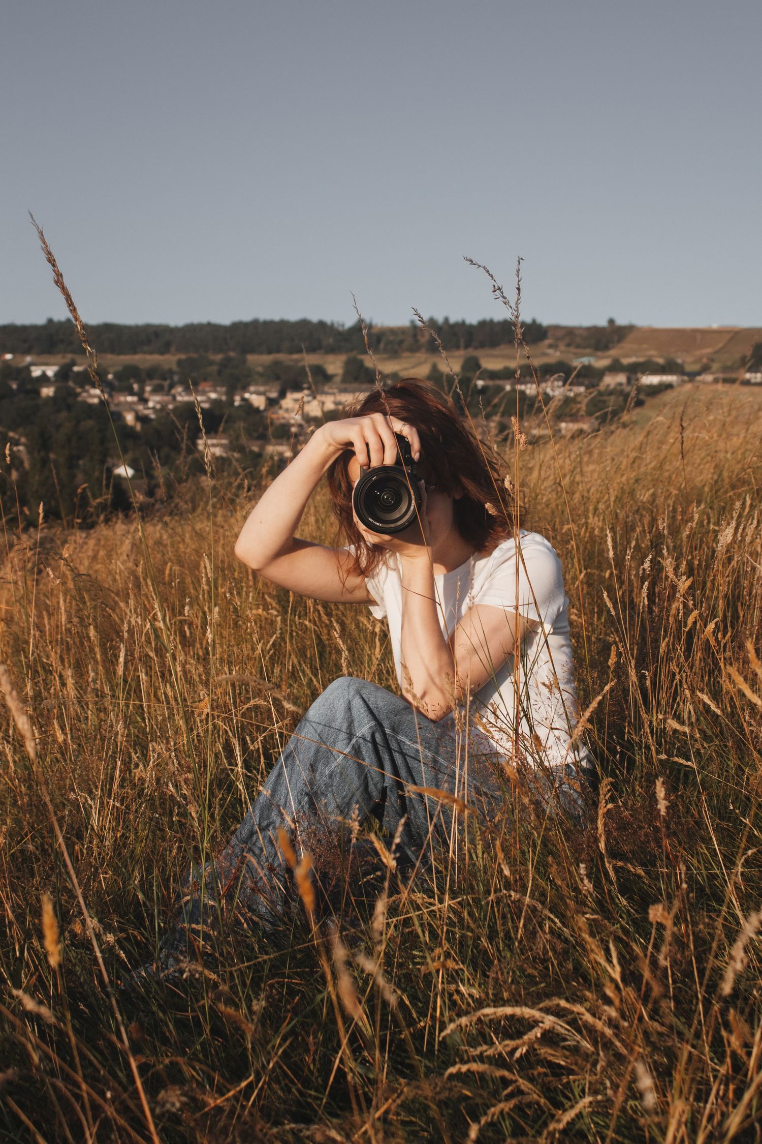 Woman sitting in a golden field photographing with a mirrorless camera at sunset.