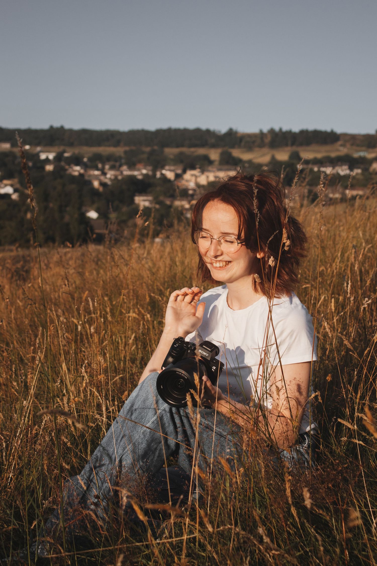 A smiling young woman with glasses sits in tall golden grass at sunset, holding a camera.
