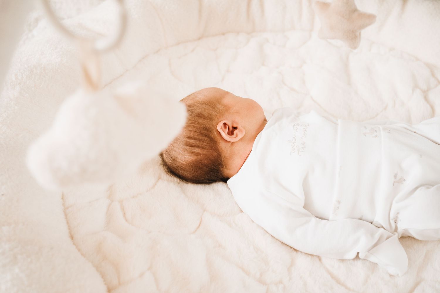 Newborn baby lying on a white quilted surface in a white onesie, viewed from above.