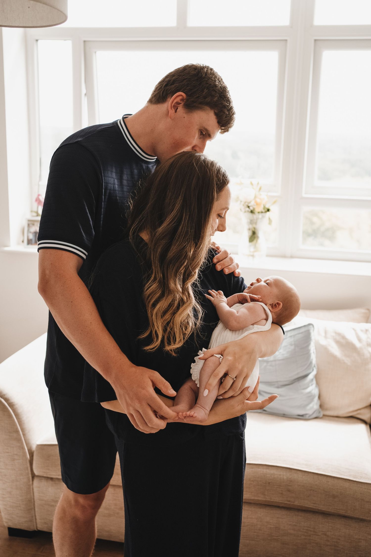 Young couple in black outfits lovingly holding their newborn baby in a bright living room.