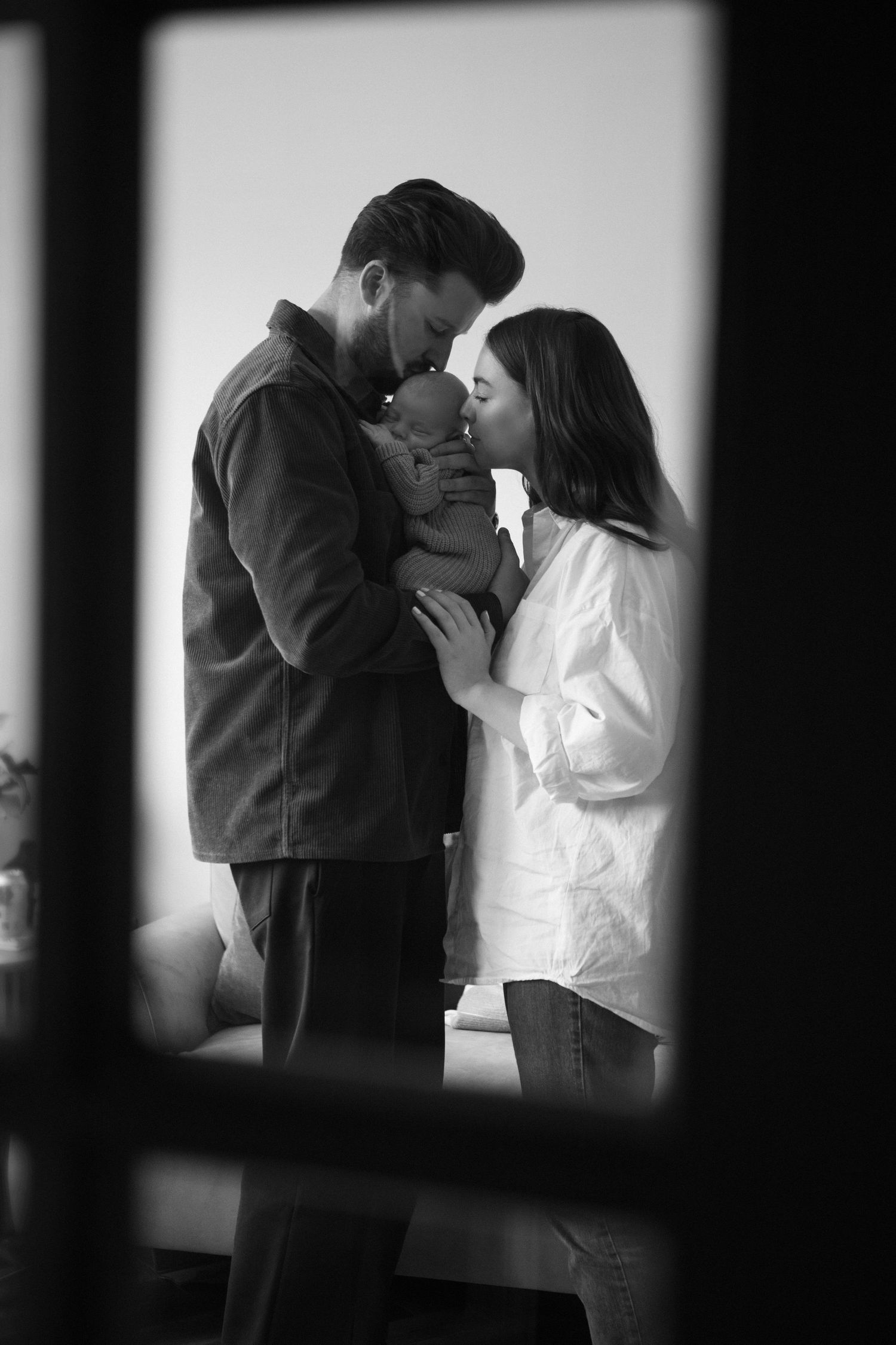 Black and white photo of parents kissing their newborn baby together in a loving embrace.
