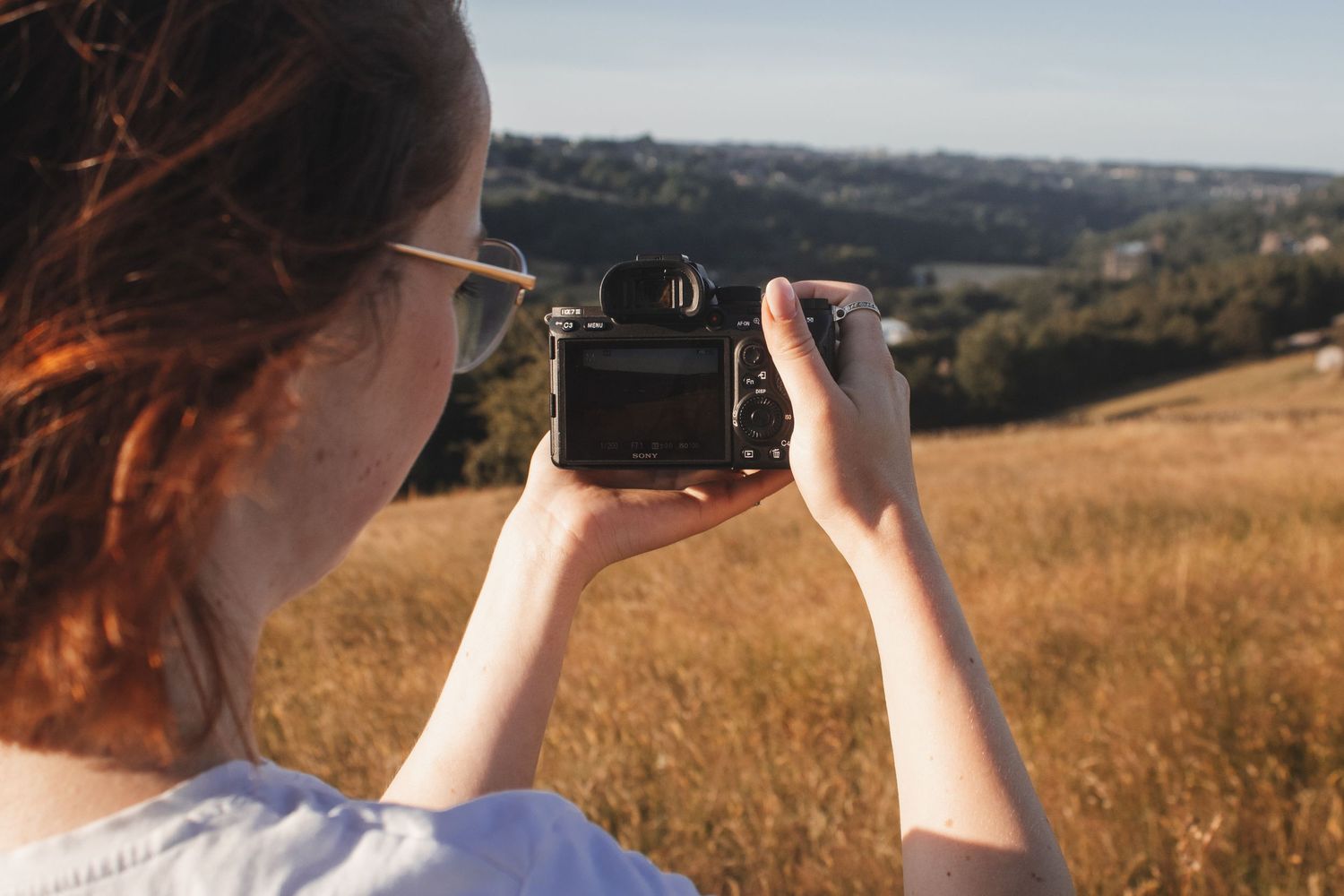 Redhead woman photographing golden field landscape with Sony camera at golden hour.