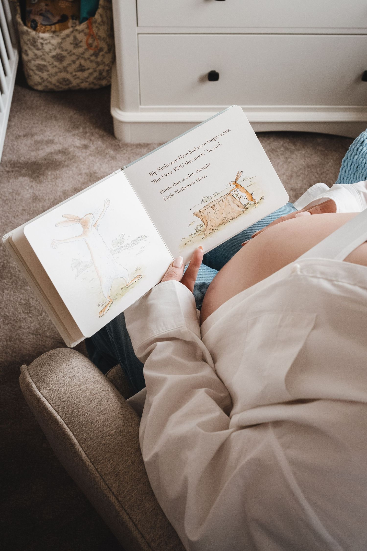 A pregnant woman relaxes in a chair reading a illustrated children's storybook to her unborn baby.