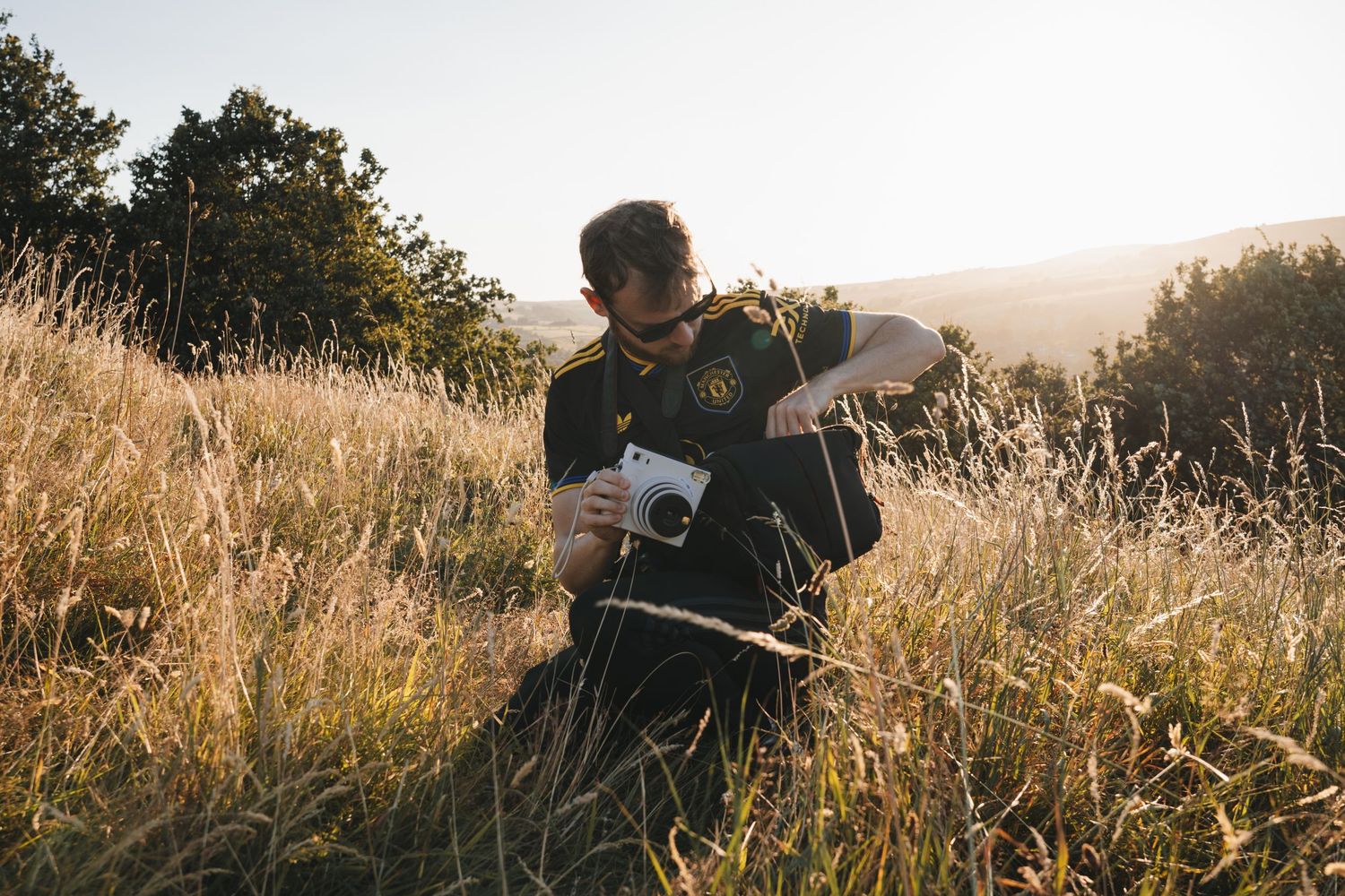 A man sits in a sunlit meadow reviewing photos on a camera surrounded by golden grass and trees.