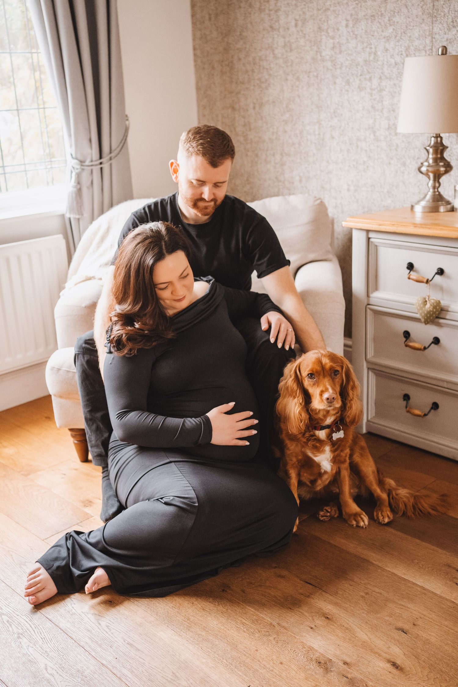A couple and their golden Cocker Spaniel sit together on a wooden floor in a cozy living room.