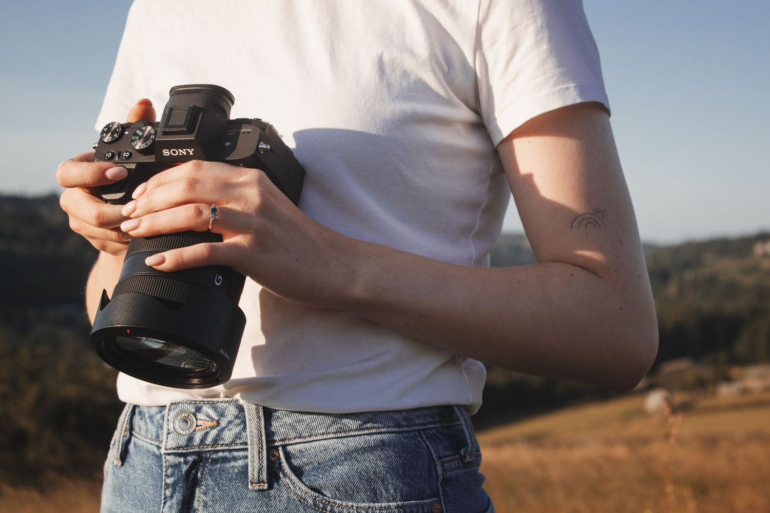 Woman in white t-shirt holding Sony mirrorless camera in sunny outdoor field setting.