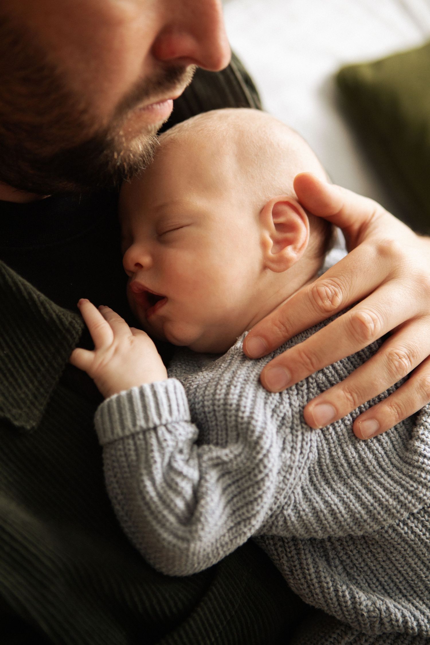 Father gently kissing his sleeping newborn baby dressed in a soft grey knit outfit.