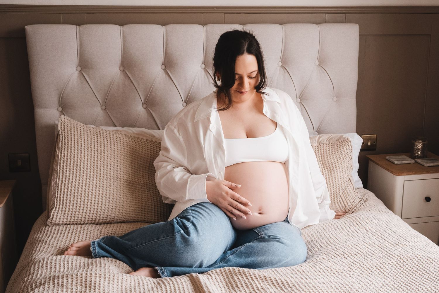 Pregnant dark-haired woman in white shirt and jeans sitting on bed cradling her baby bump.