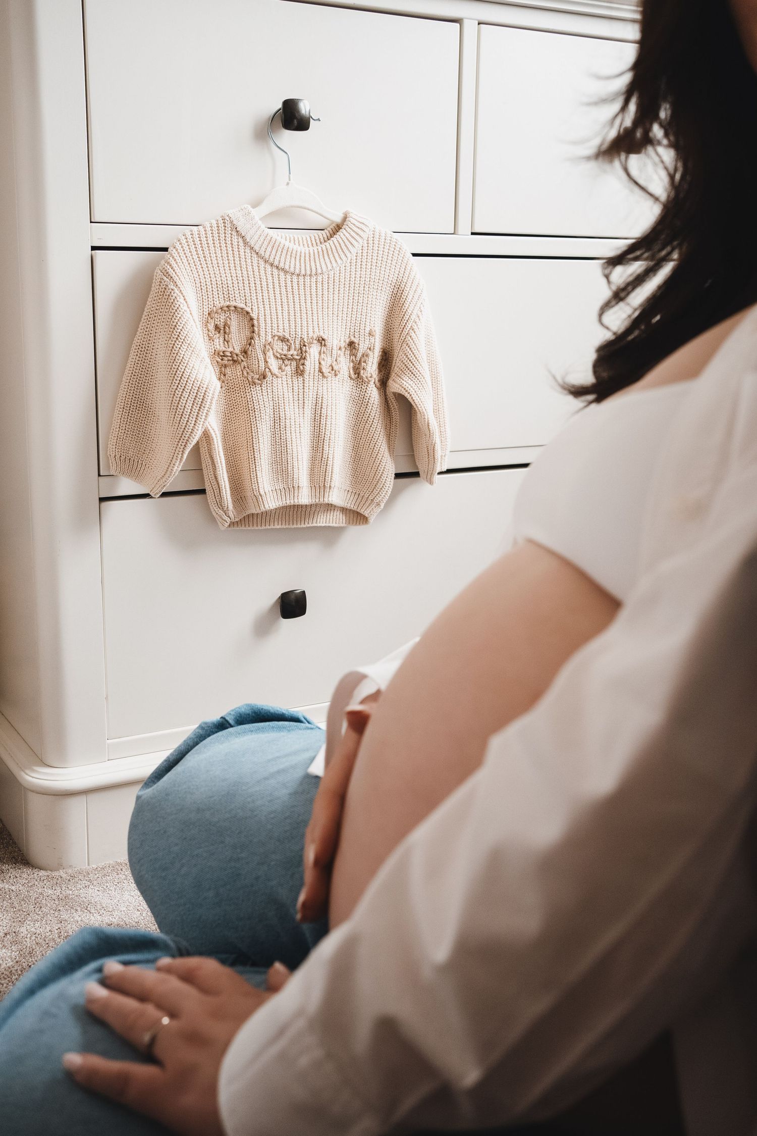 Pregnant woman sitting on floor holding baby bump, looking at personalized cream knit sweater hanging on white dresser.