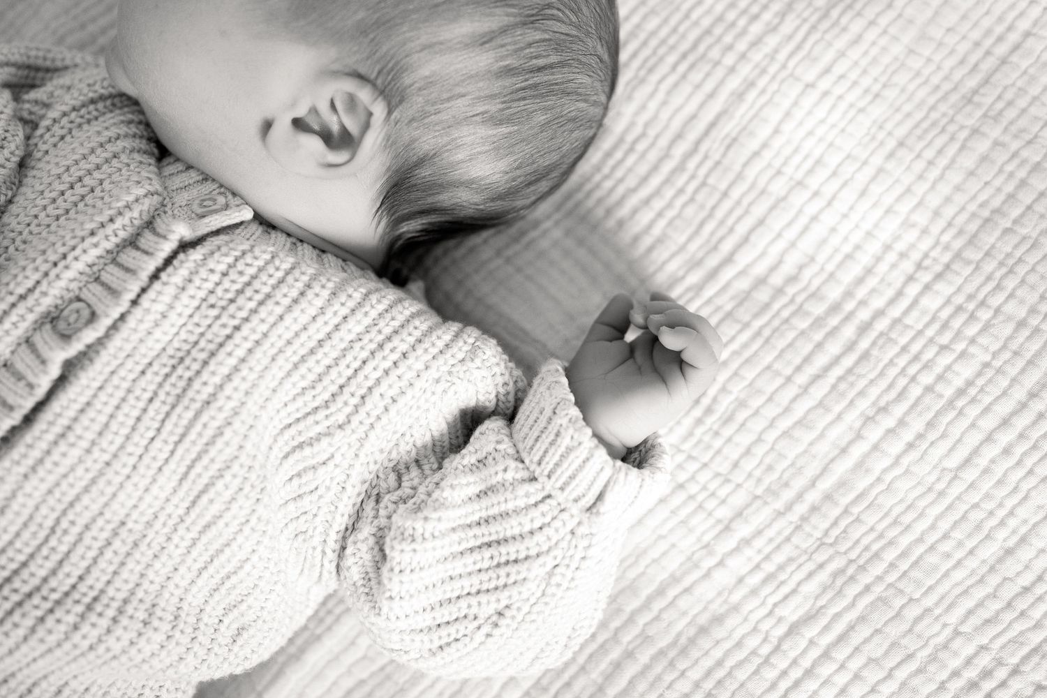 Black and white photo of a sleeping newborn baby in a knit sweater lying on a soft blanket.