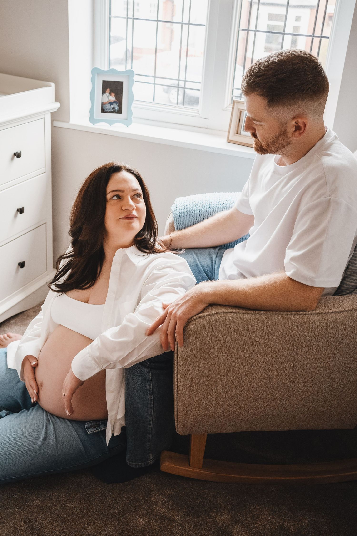 A pregnant woman sits on the floor while her partner sits in a rocking chair beside her in a nursery.