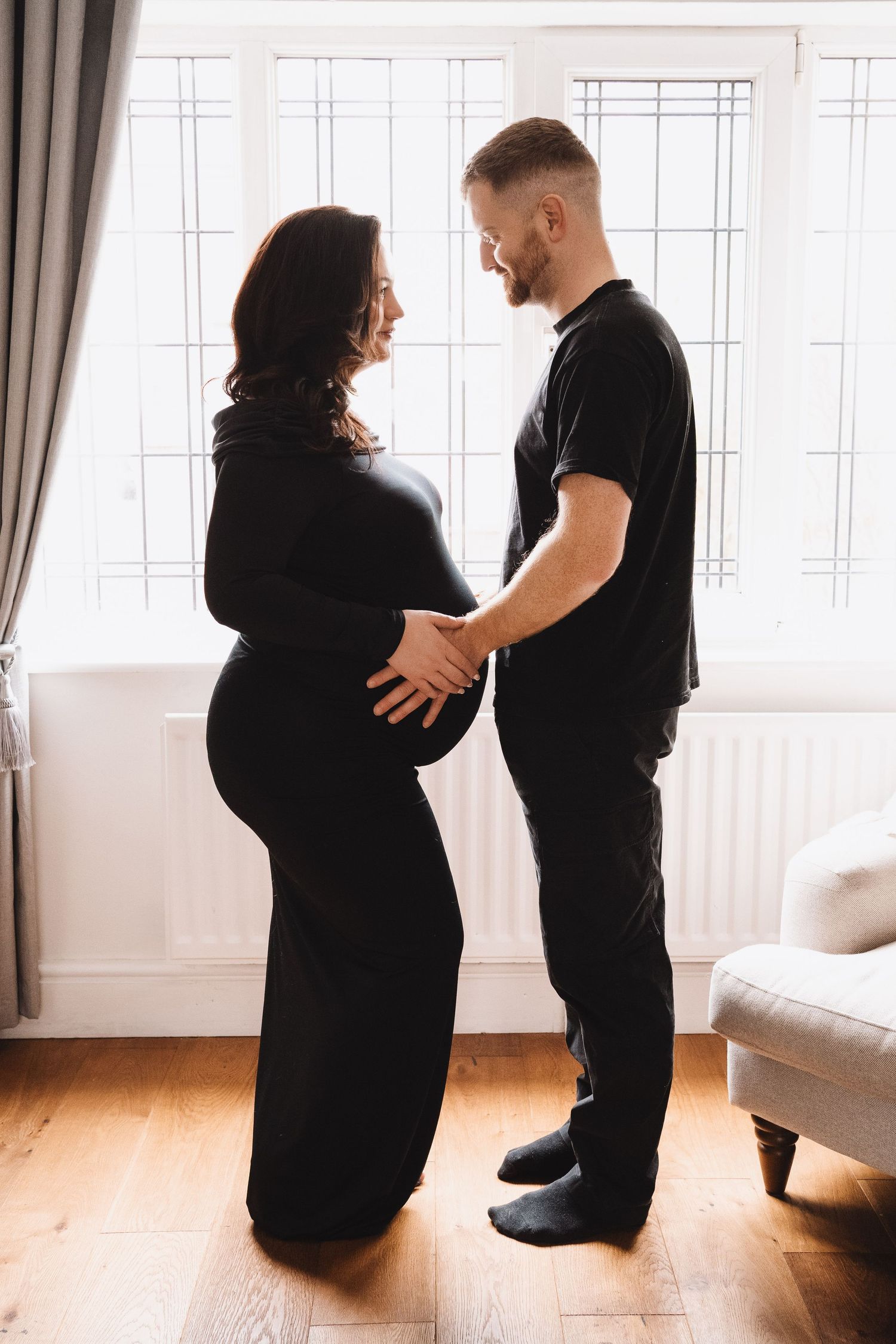 A couple dressed in black stands by a bright window, smiling as the man holds the pregnant belly.