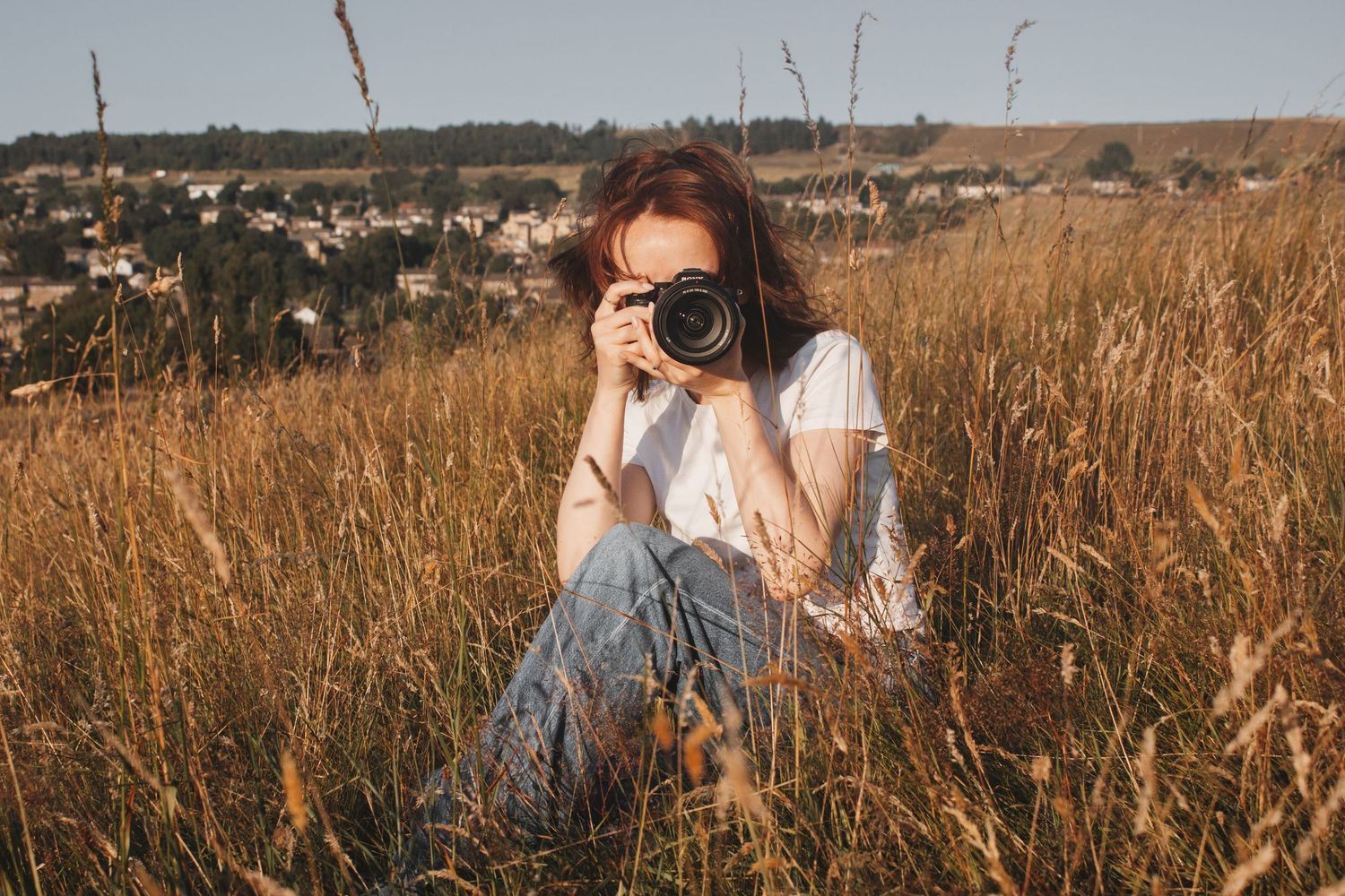 Young woman with red hair photographing with a camera while sitting in a field.