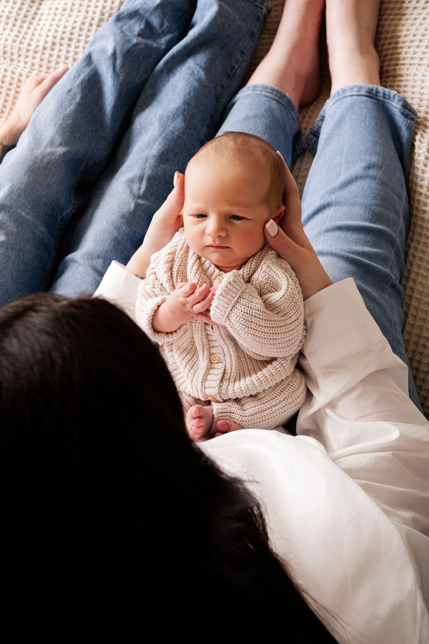 Overhead view of a newborn baby in a beige knit outfit lying between parents' legs on a soft surface.