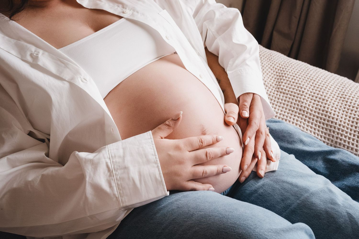 Close-up of a pregnant womans baby bump with hands gently cradling it while sitting on sofa.