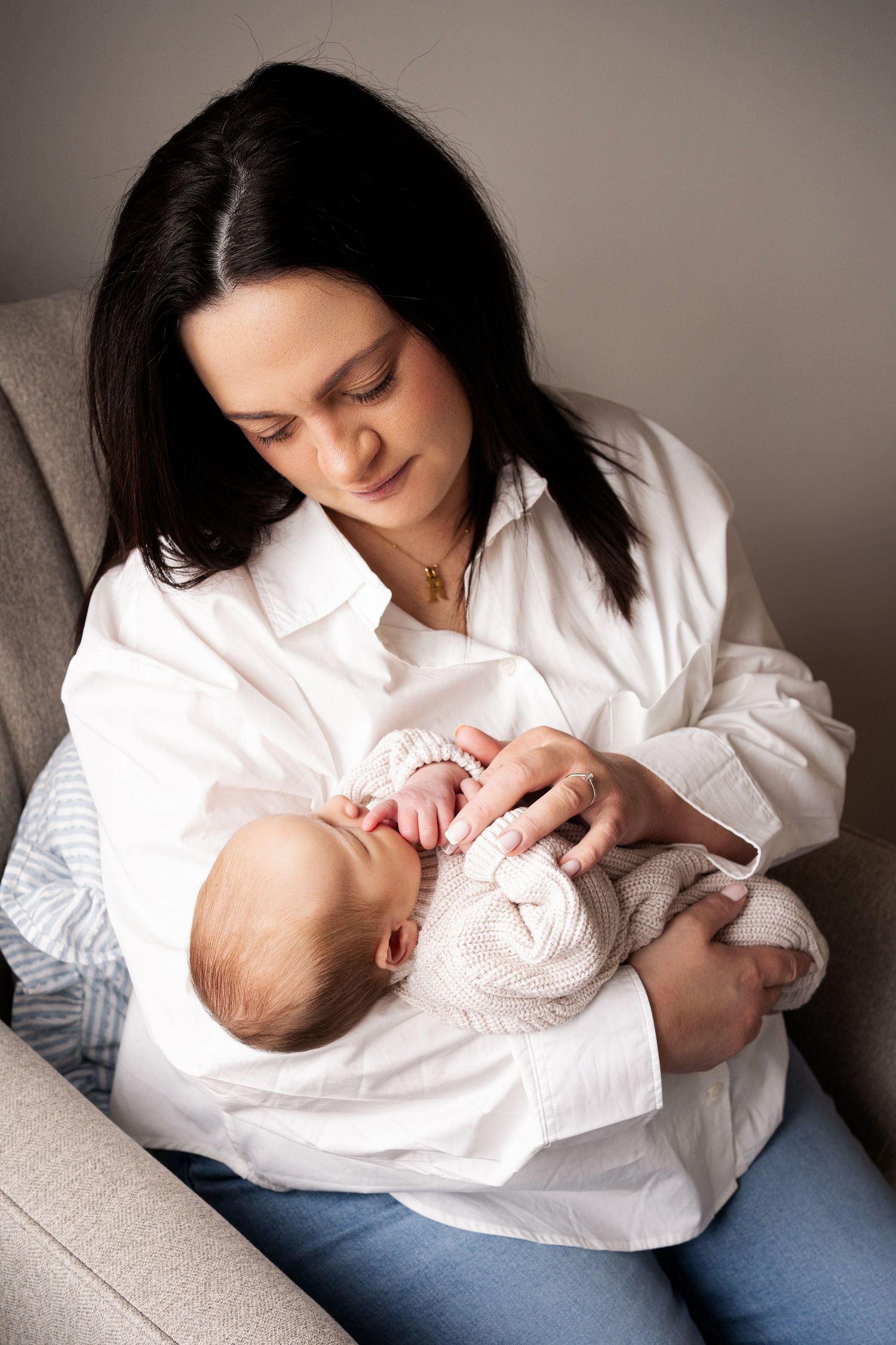 A dark-haired mother in a white shirt tenderly holds her newborn baby while sitting in a gray armchair.