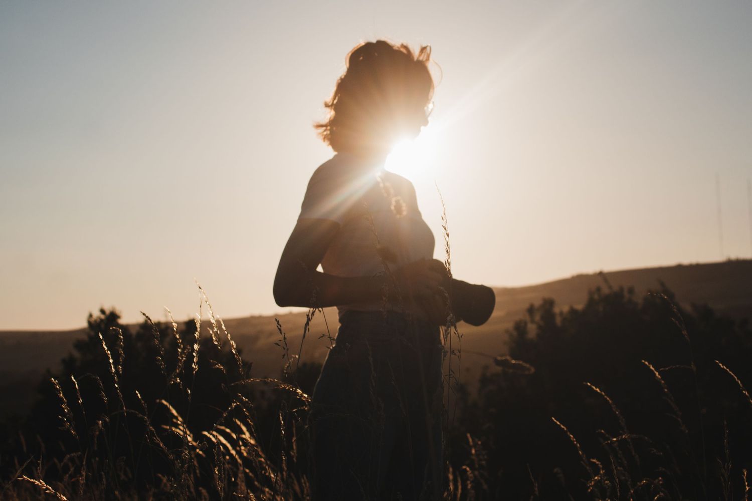A silhouetted woman stands in a sunlit field holding a camera with warm golden light behind her.