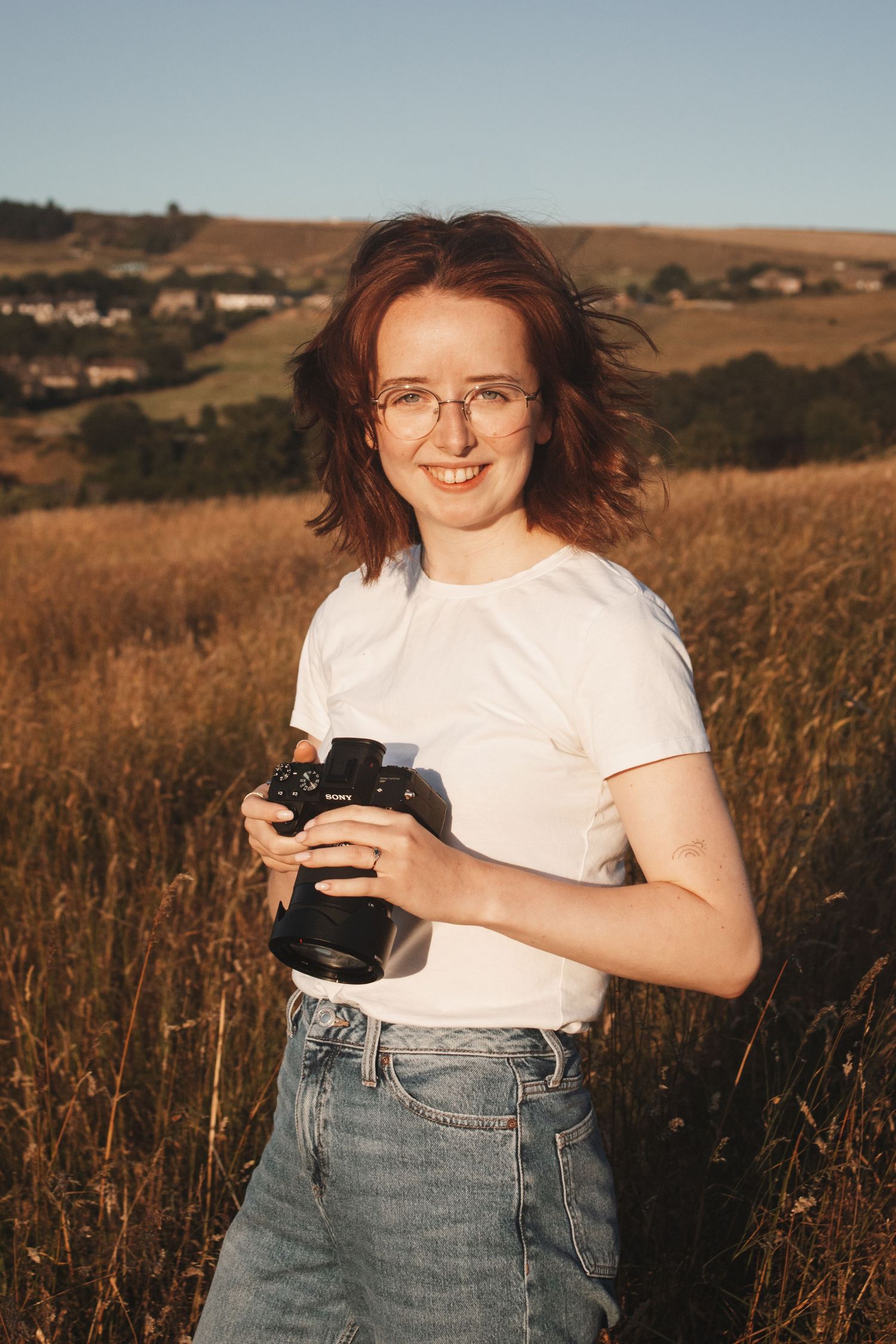 A smiling young woman with red hair and glasses holds a Sony camera in a golden sunlit field.