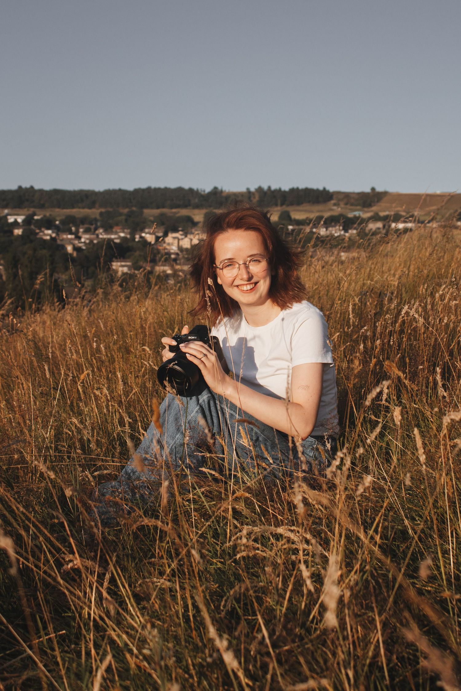 Happy redhead woman with glasses holding a camera while sitting in a sunny field.