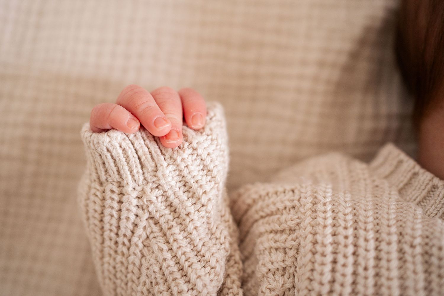Close-up of a newborn baby's tiny hand gripping the sleeve of a cozy beige knit sweater.