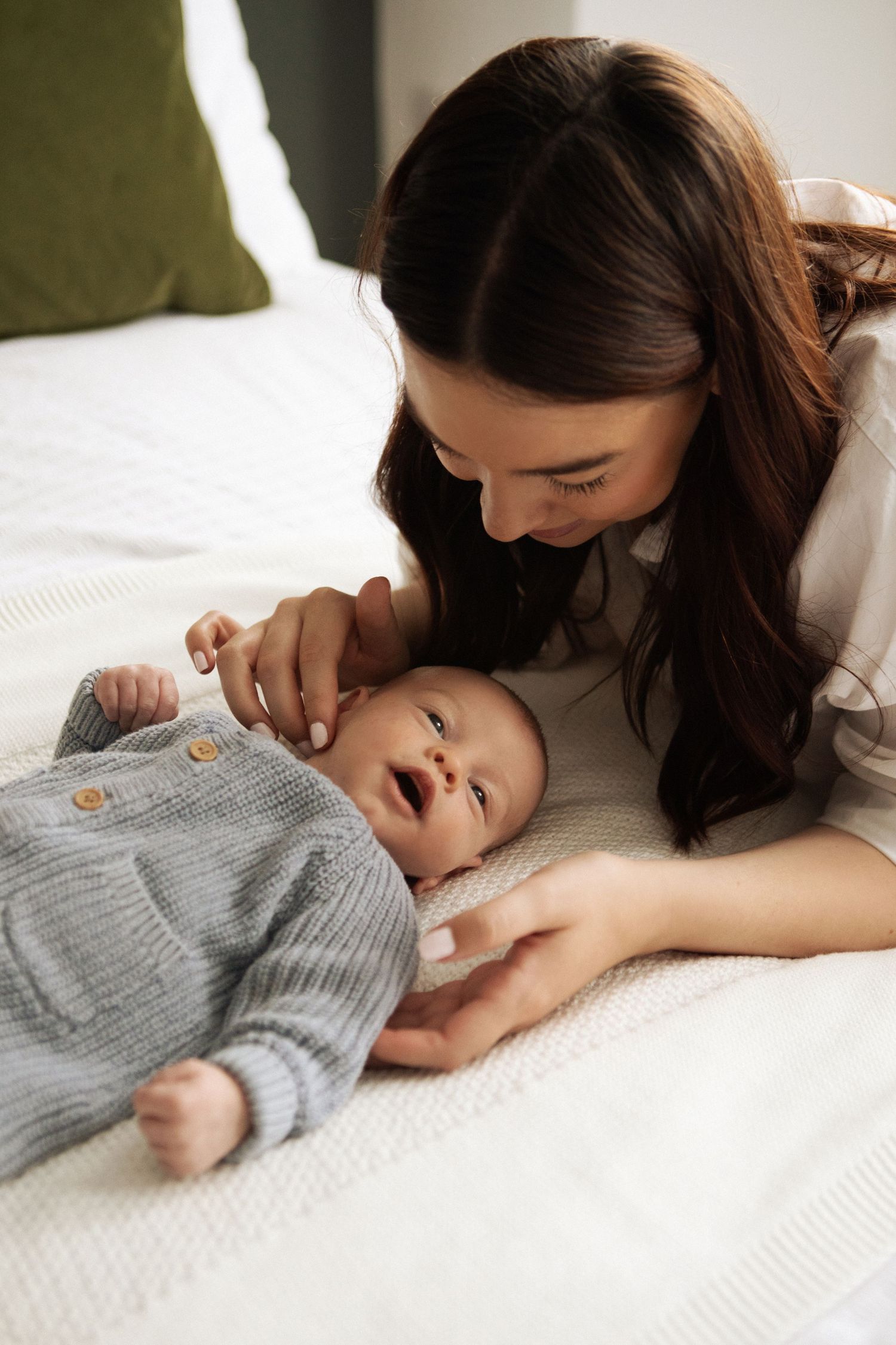 A smiling dark-haired mother leans over her happy newborn baby dressed in a grey knit romper.