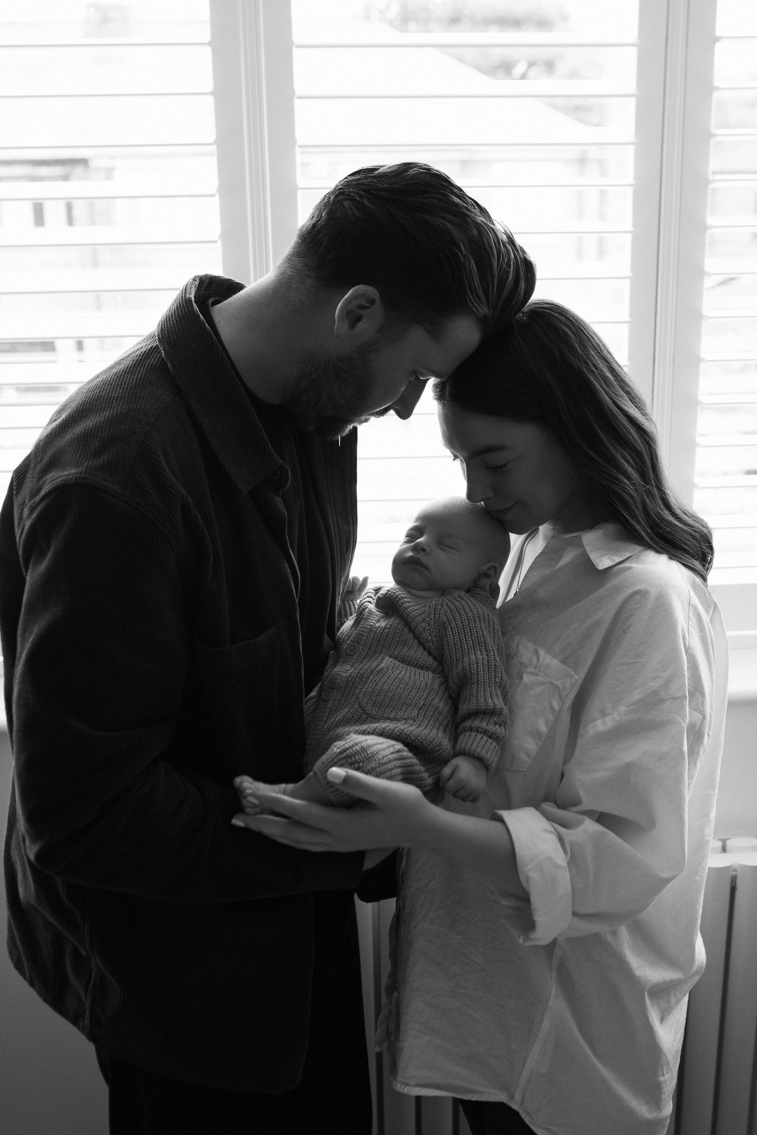 Parents holding newborn baby together by window in black and white.