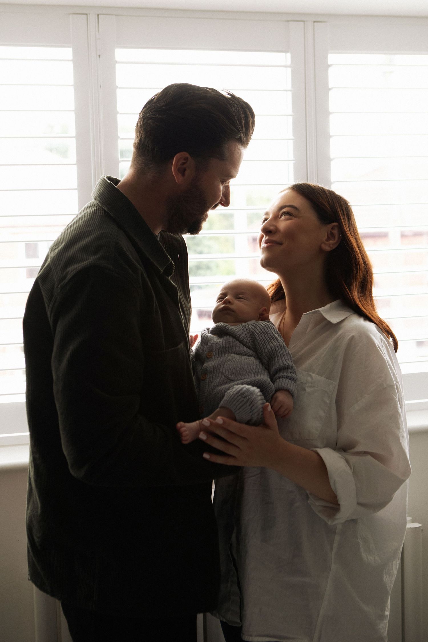 Happy couple standing by window holding their newborn baby, sharing a tender moment together.