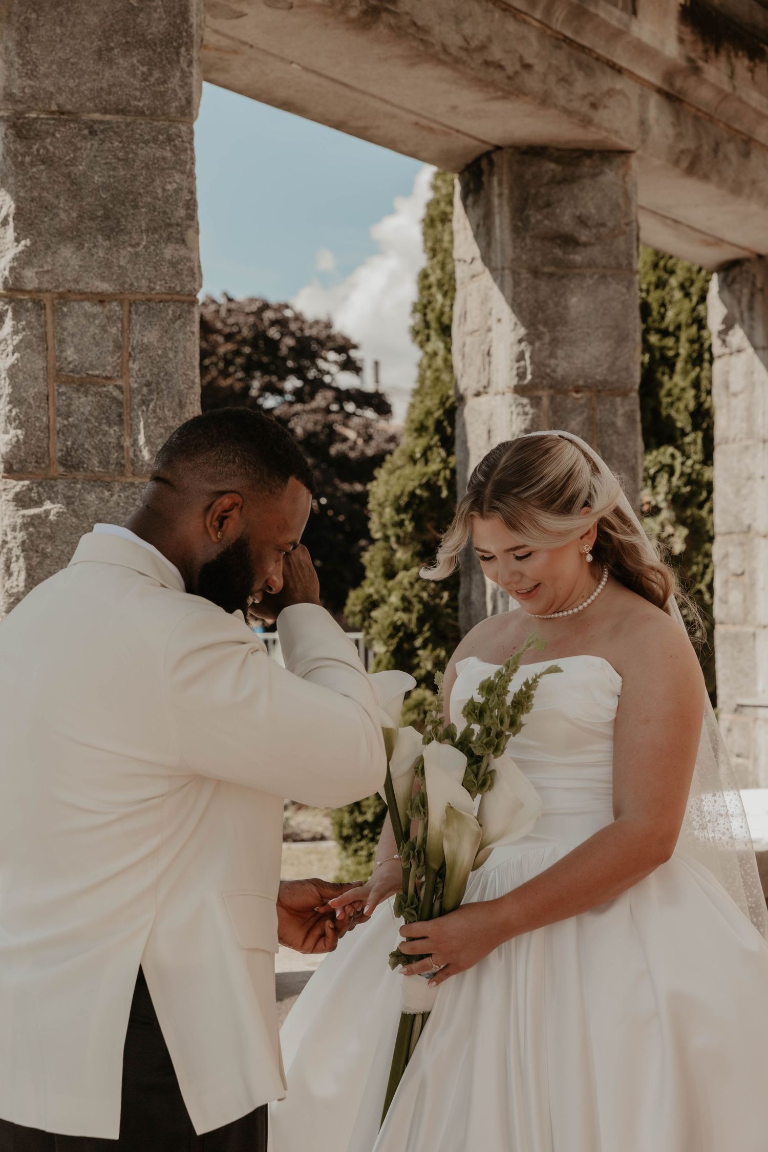 Emotional groom in white suit wiping tears beside bride in strapless wedding gown holding calla lilies at stone venue.