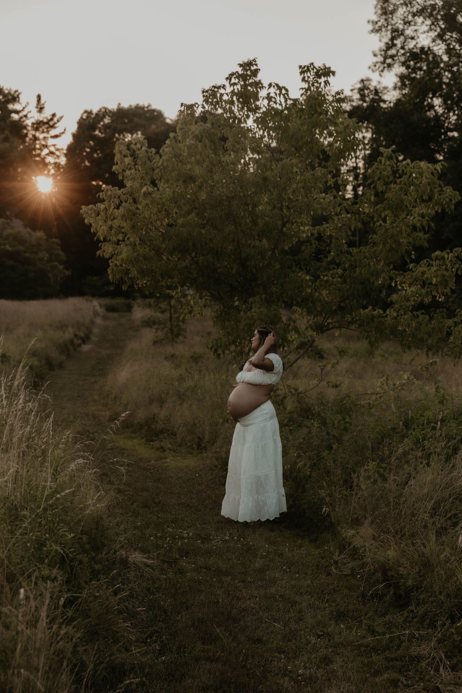Pregnant woman in white dress standing on a garden path at golden hour sunset surrounded by lush green trees.