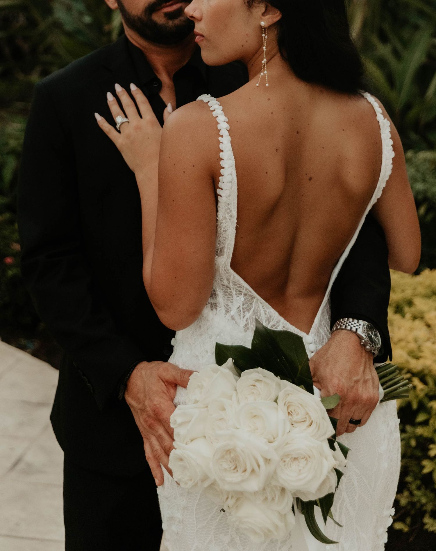 Bride in backless lace gown holds white rose bouquet embraced by groom in black suit at outdoor wedding.