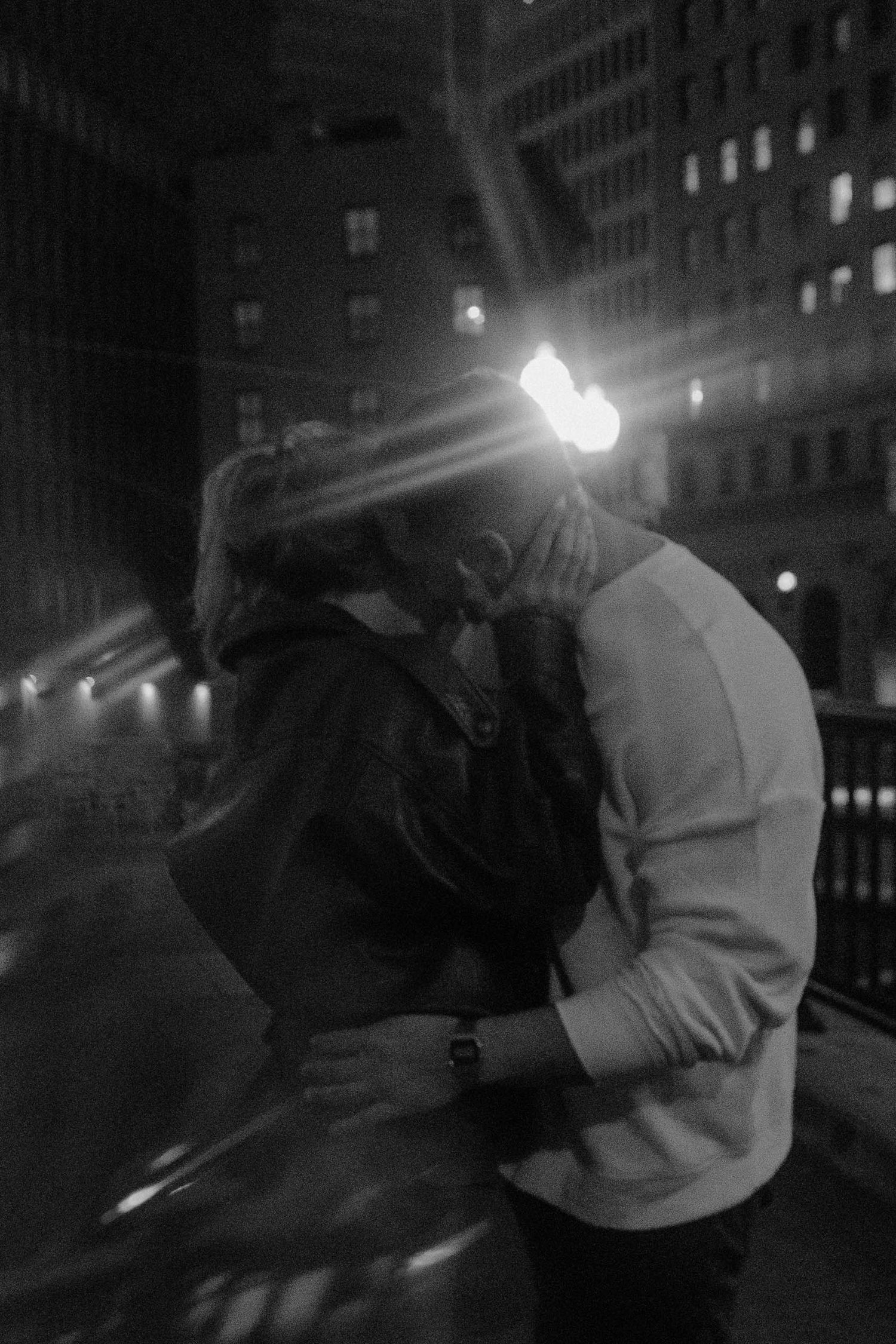 Black and white photo of a couple kissing at night in the city with bright light flare behind them.