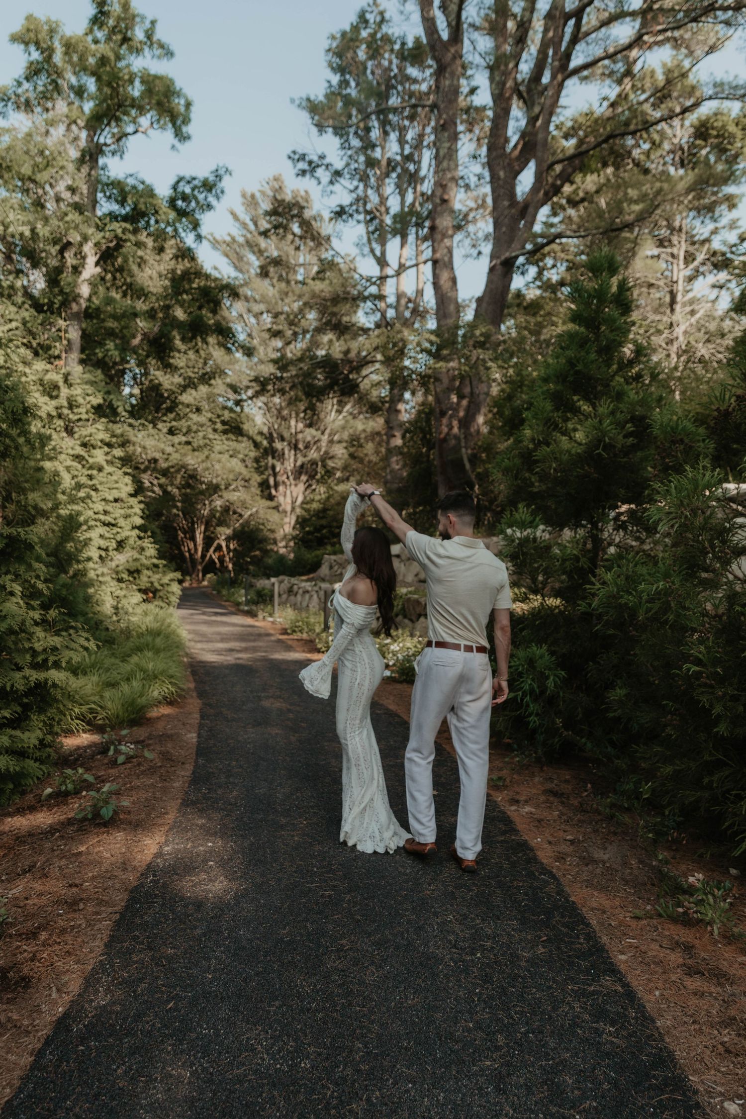 A bride and groom dance together on a garden path surrounded by lush green trees during their outdoor wedding.