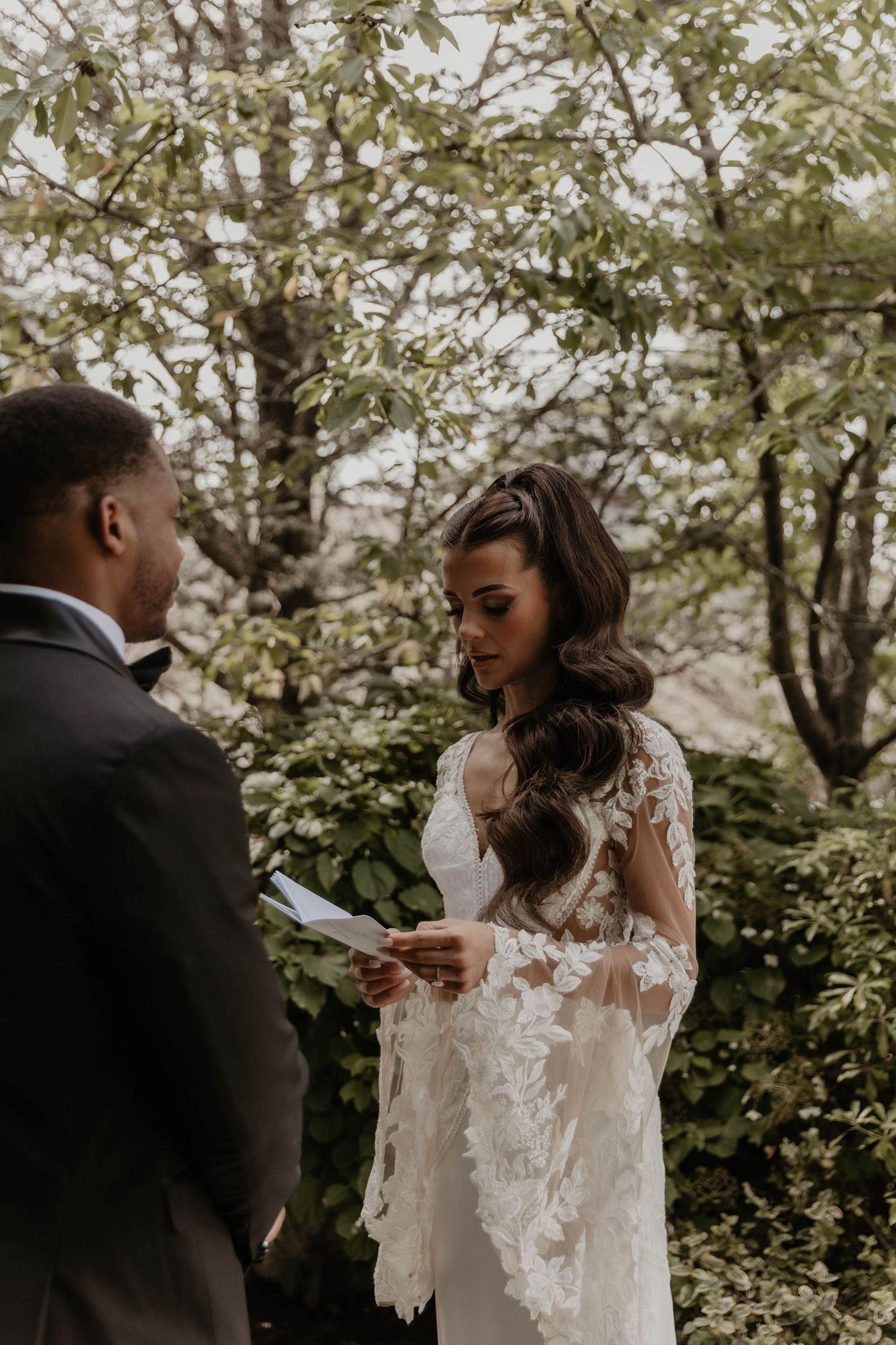 Bride in lace gown reads wedding vows to groom in suit outdoors among lush green trees during intimate ceremony.