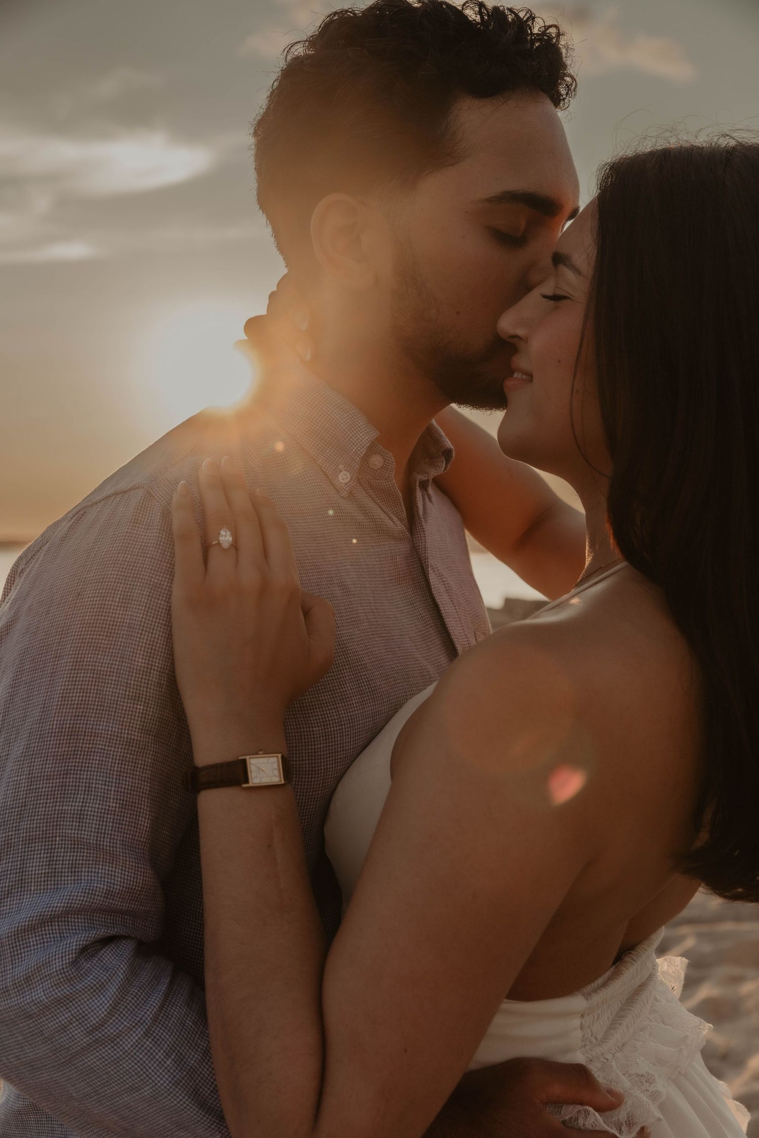 Couple sharing a romantic sunset kiss on the beach, woman wearing engagement ring and white dress.